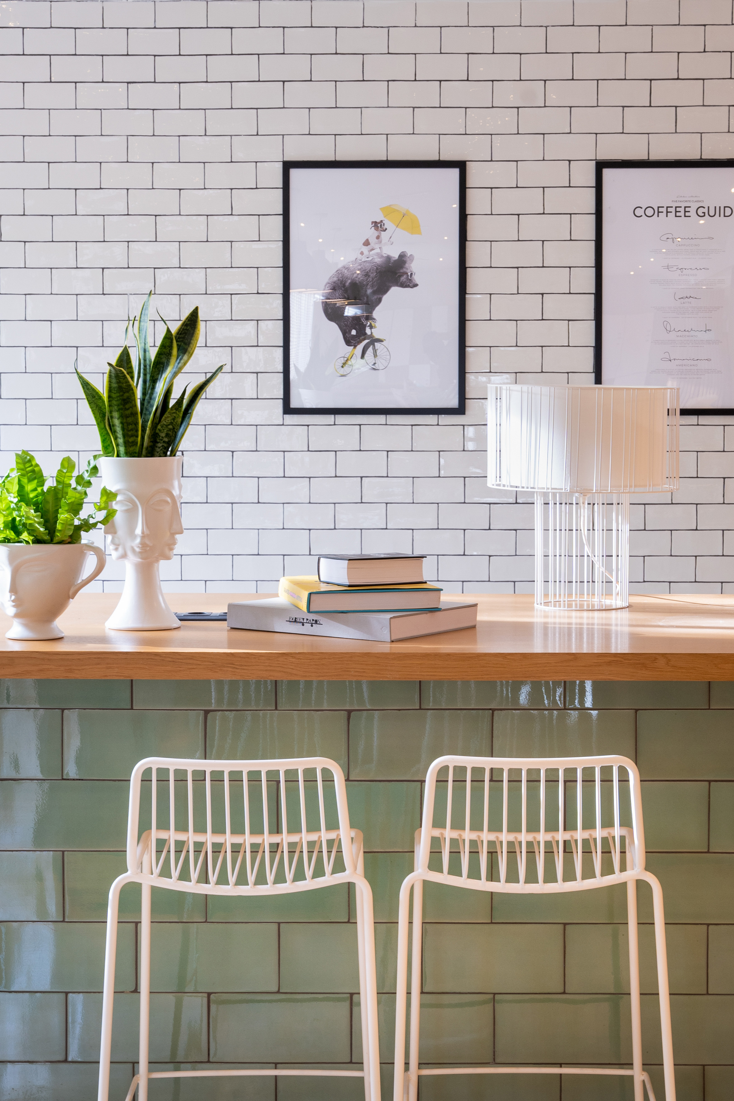 Modern cafe bar with white/green tiles, wood counter, plants, books, white stools.