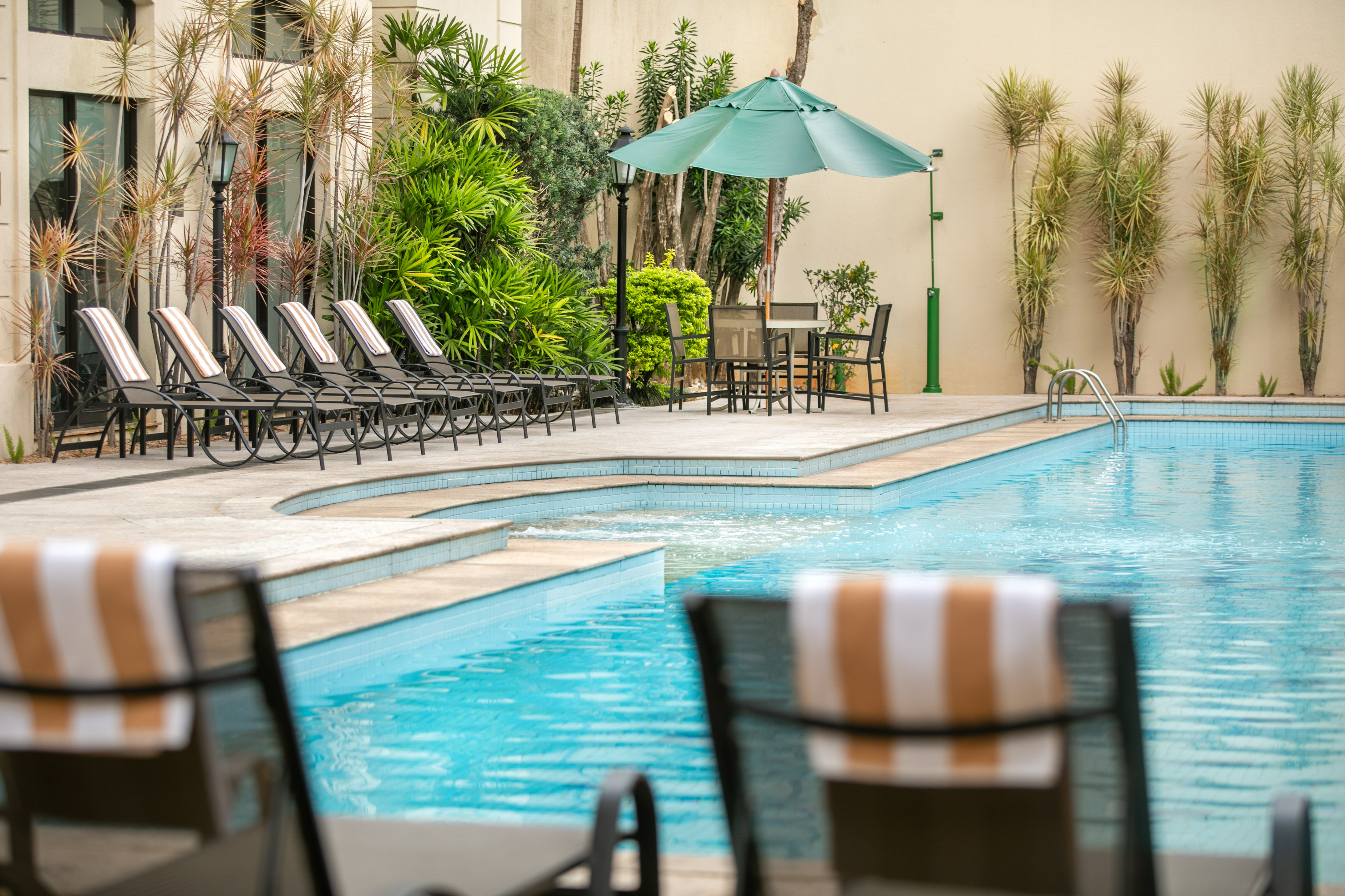 Resort swimming pool with lounge chairs, green umbrella, and tropical plants.