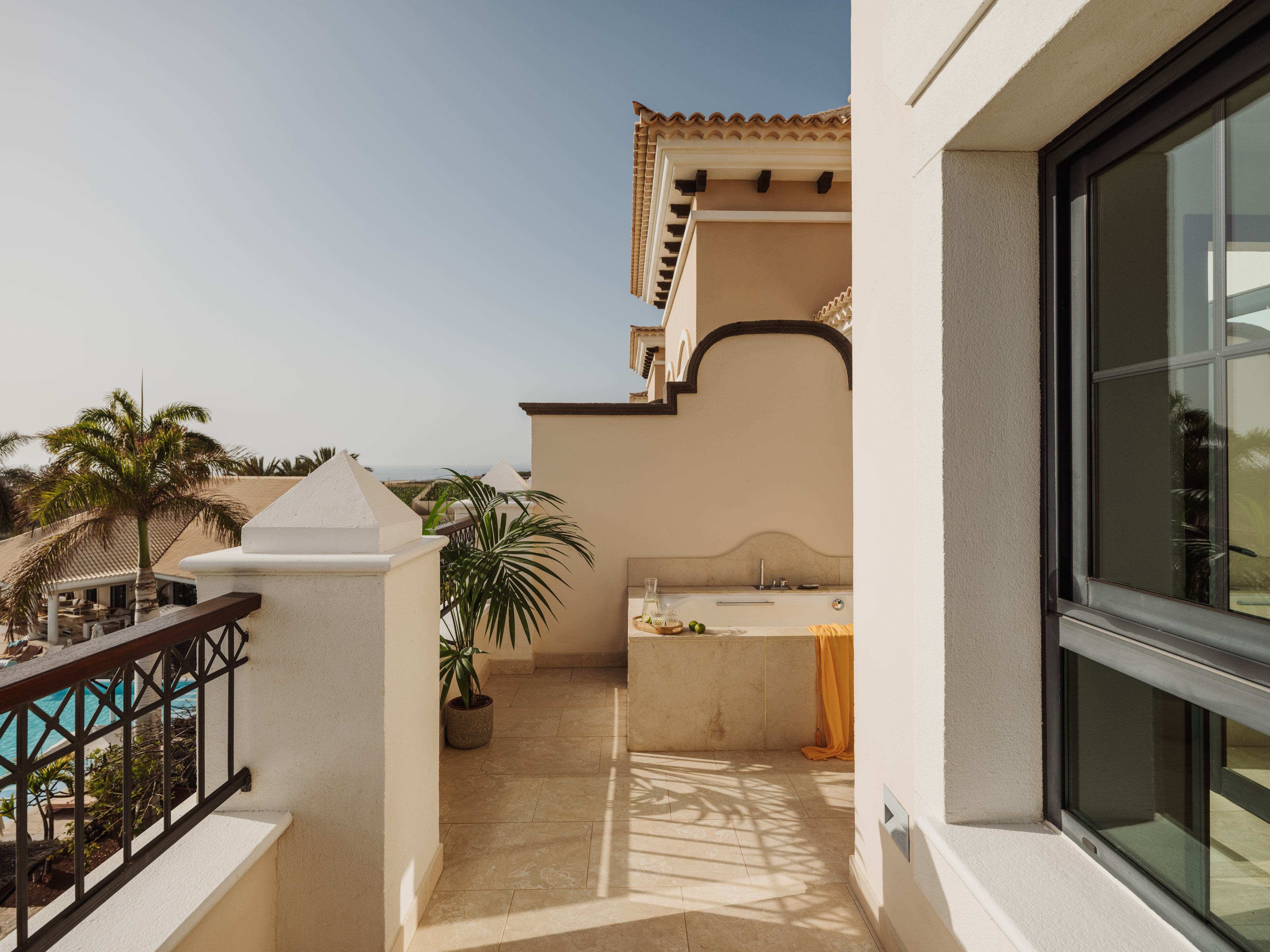 Sunny resort balcony with outdoor spa tub, palm trees, and pool view.