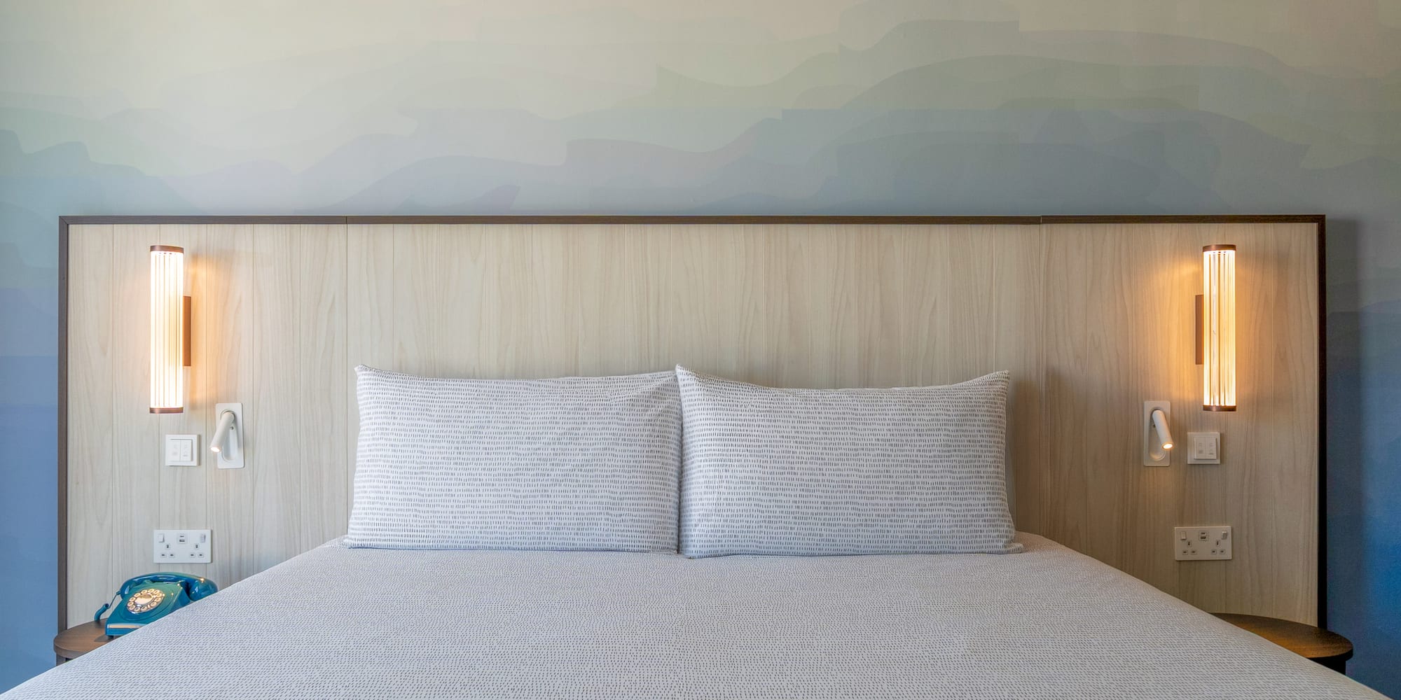 Modern hotel bedroom with bed, wooden headboard, gradient wall art, and lamps.