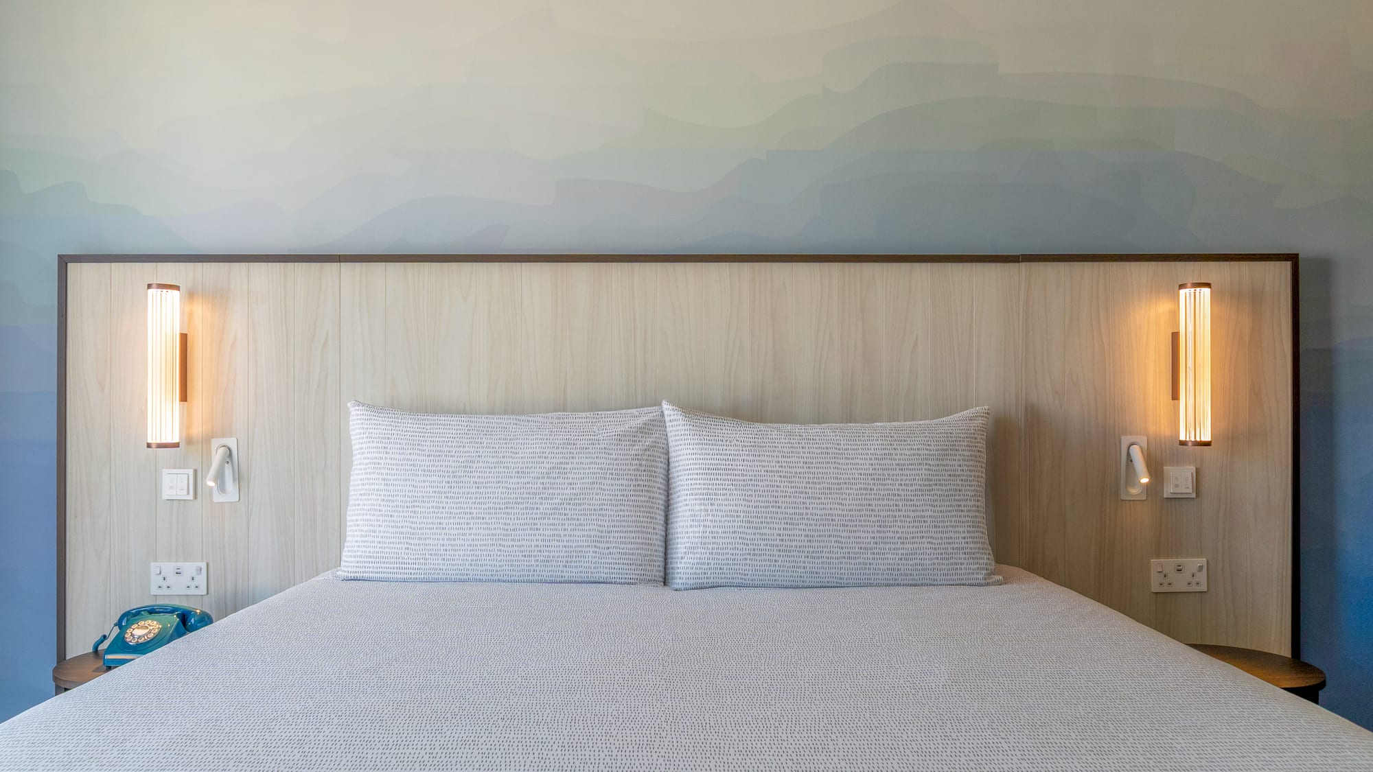 Modern hotel bedroom with bed, wooden headboard, gradient wall art, and lamps.