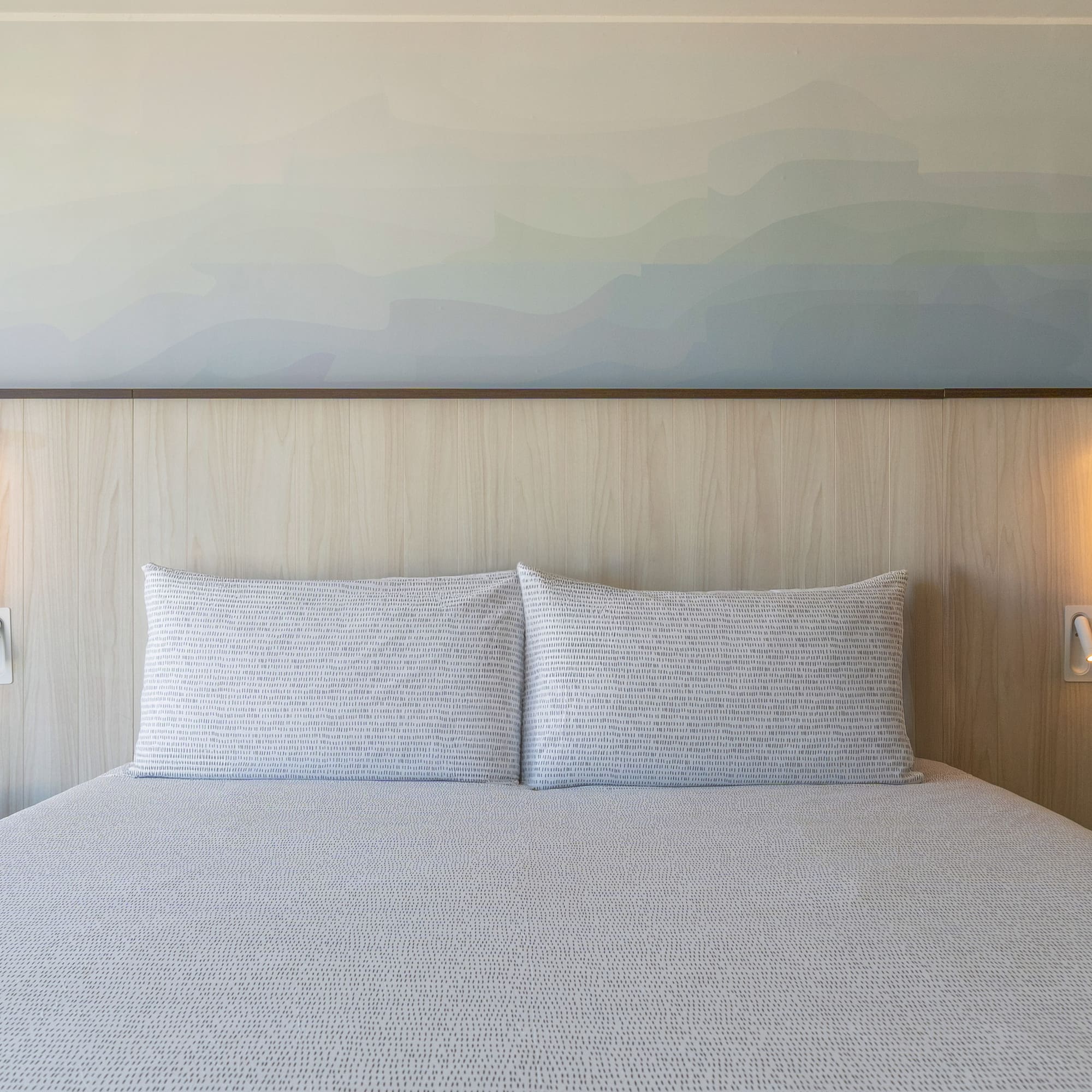 Modern hotel bedroom with bed, wooden headboard, gradient wall art, and lamps.