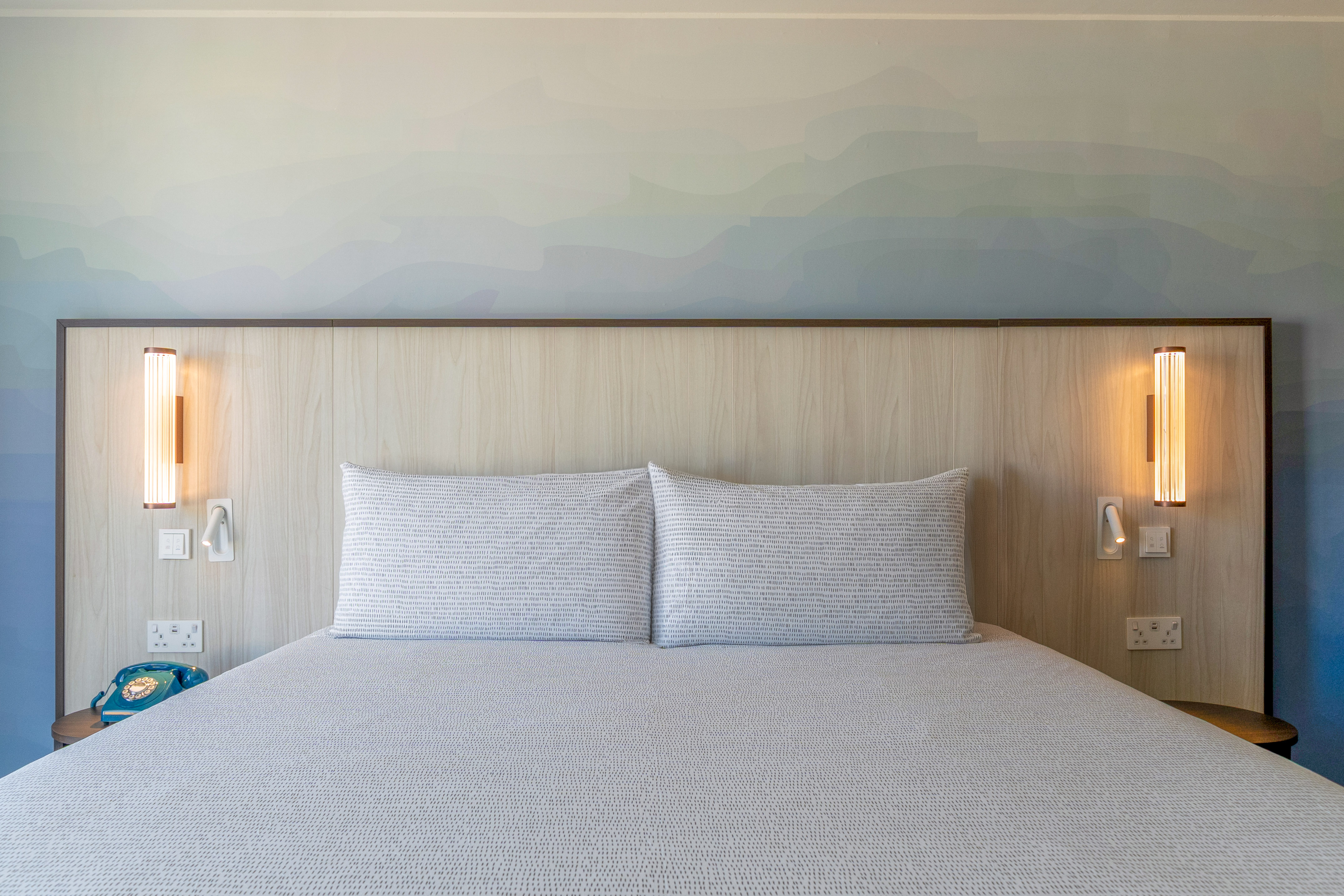 Modern hotel bedroom with bed, wooden headboard, gradient wall art, and lamps.