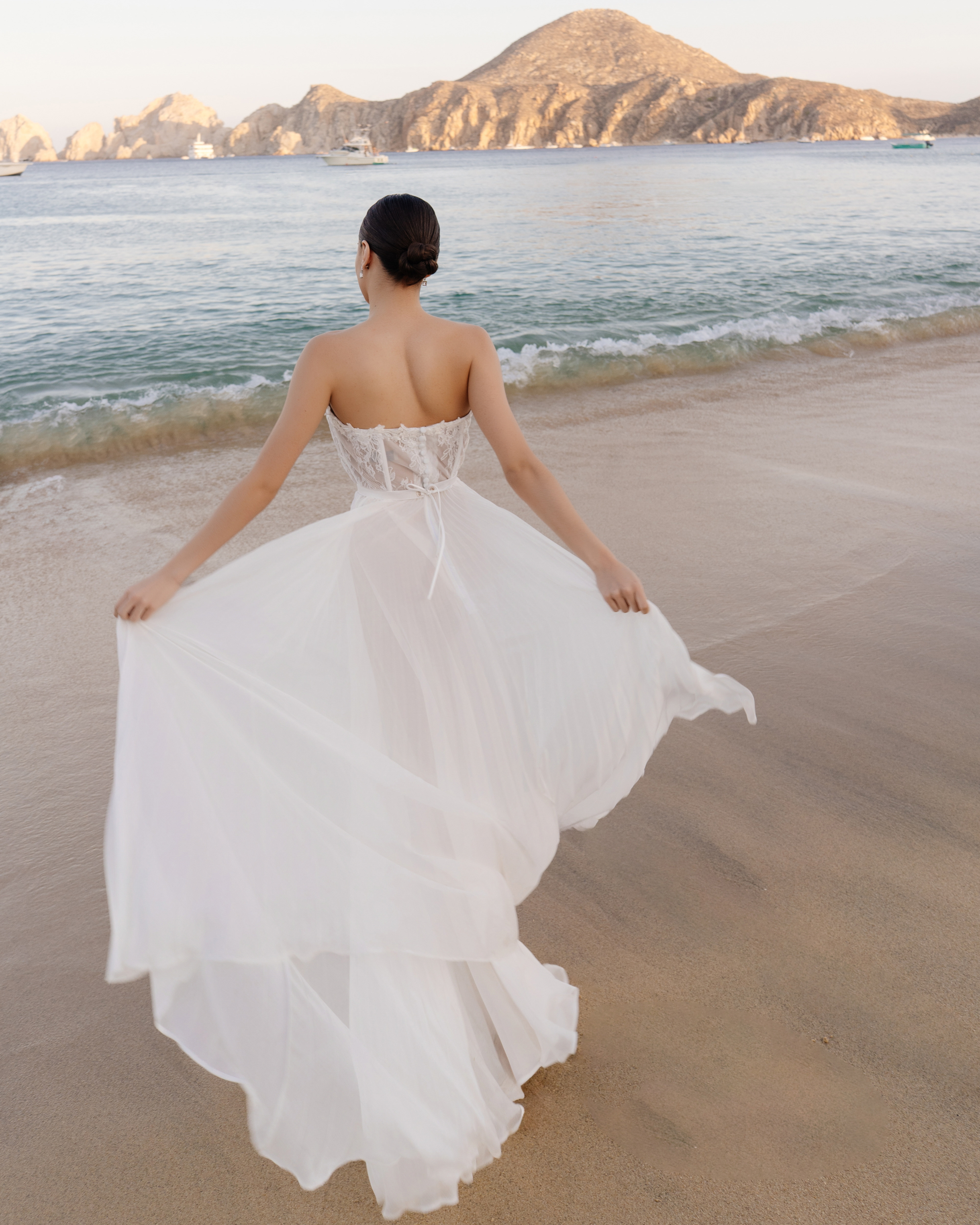 Woman in white strapless wedding dress on sandy beach, ocean, rocky islands.