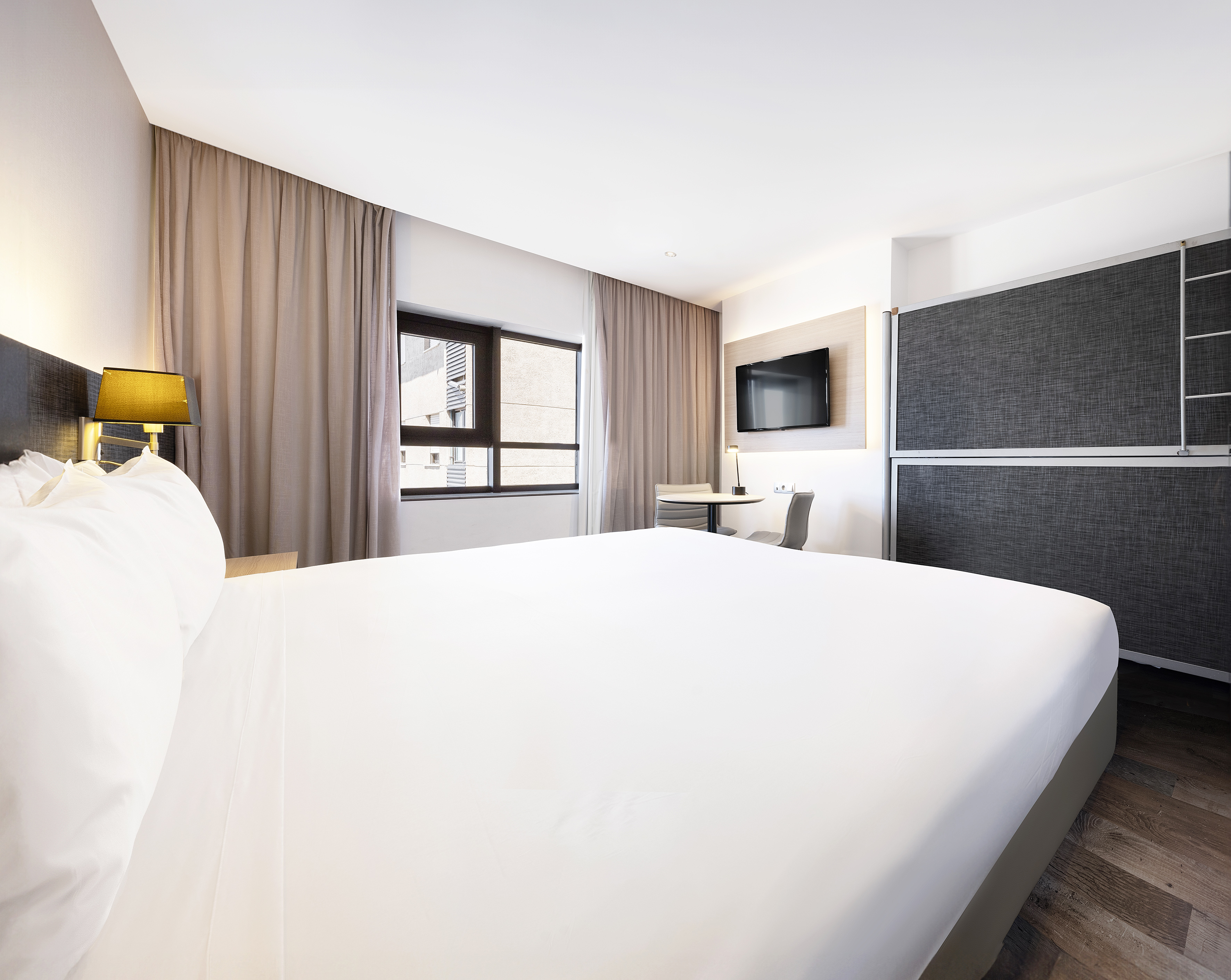 Modern hotel room with white bed, wall-mounted TV, window, and seating area.