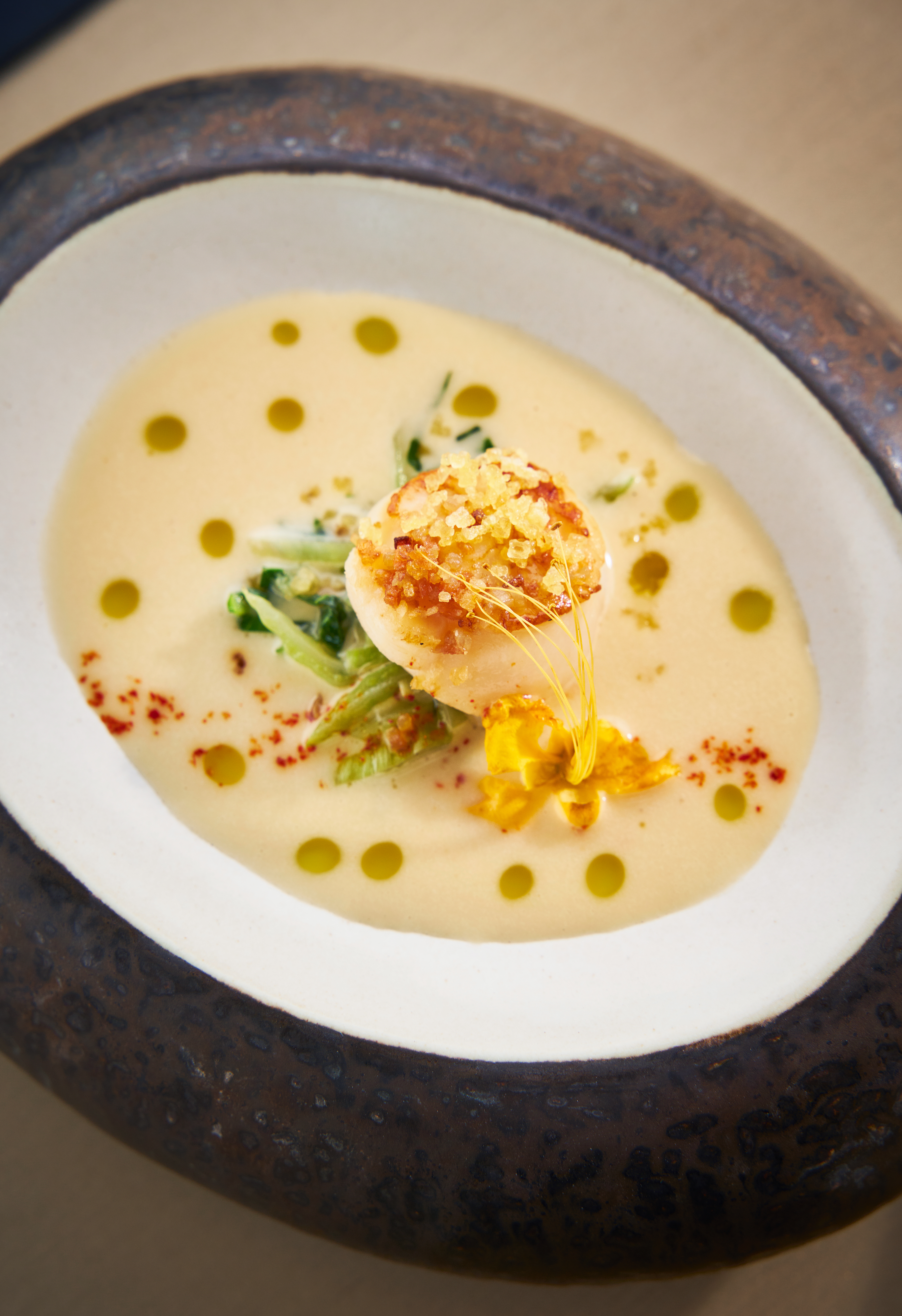 Seared scallop in creamy bisque, crispy topping, olive oil, and delicate green garnish.