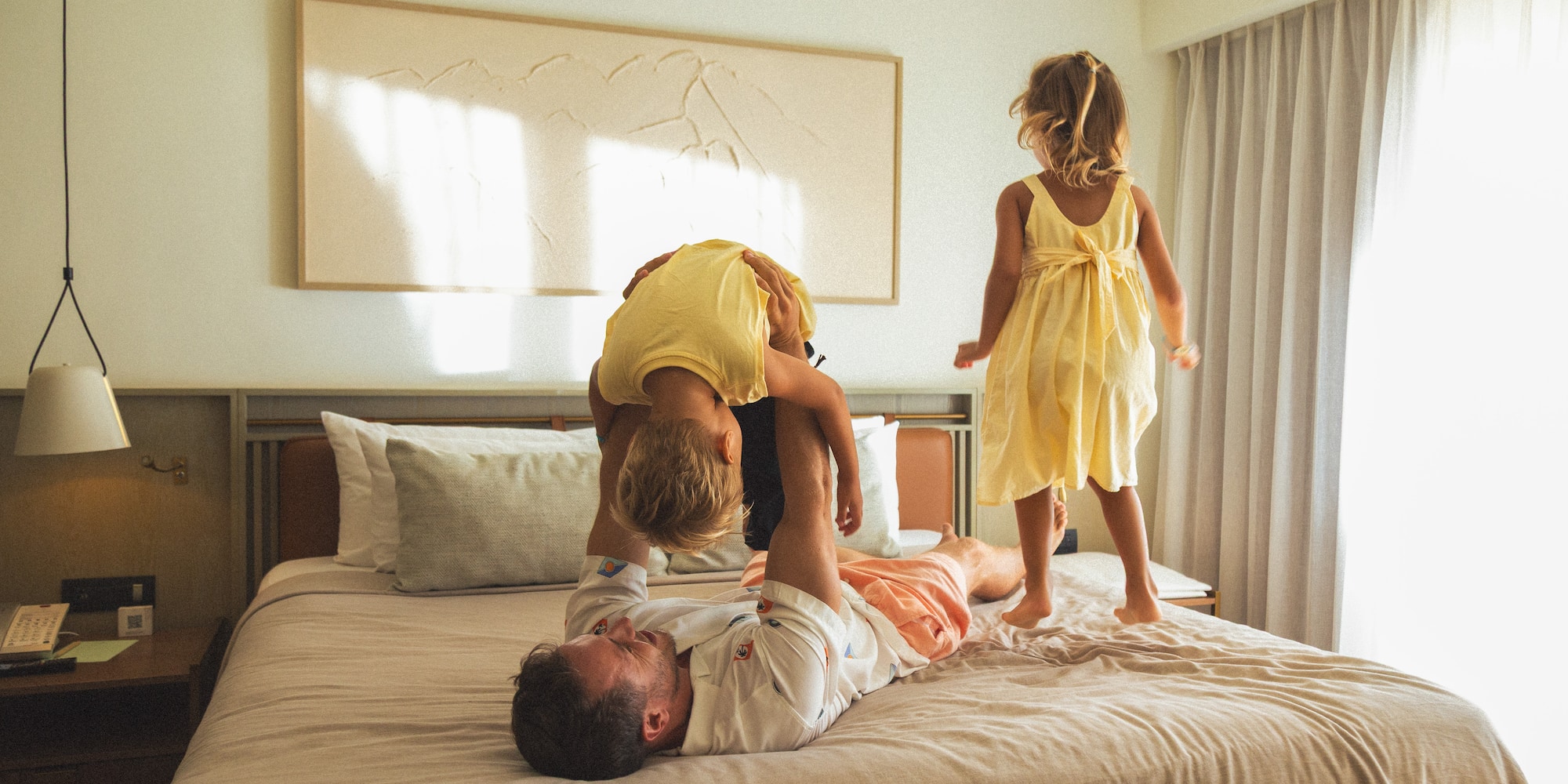 Playful dad with two kids on bed; one jumping, one held upside down.