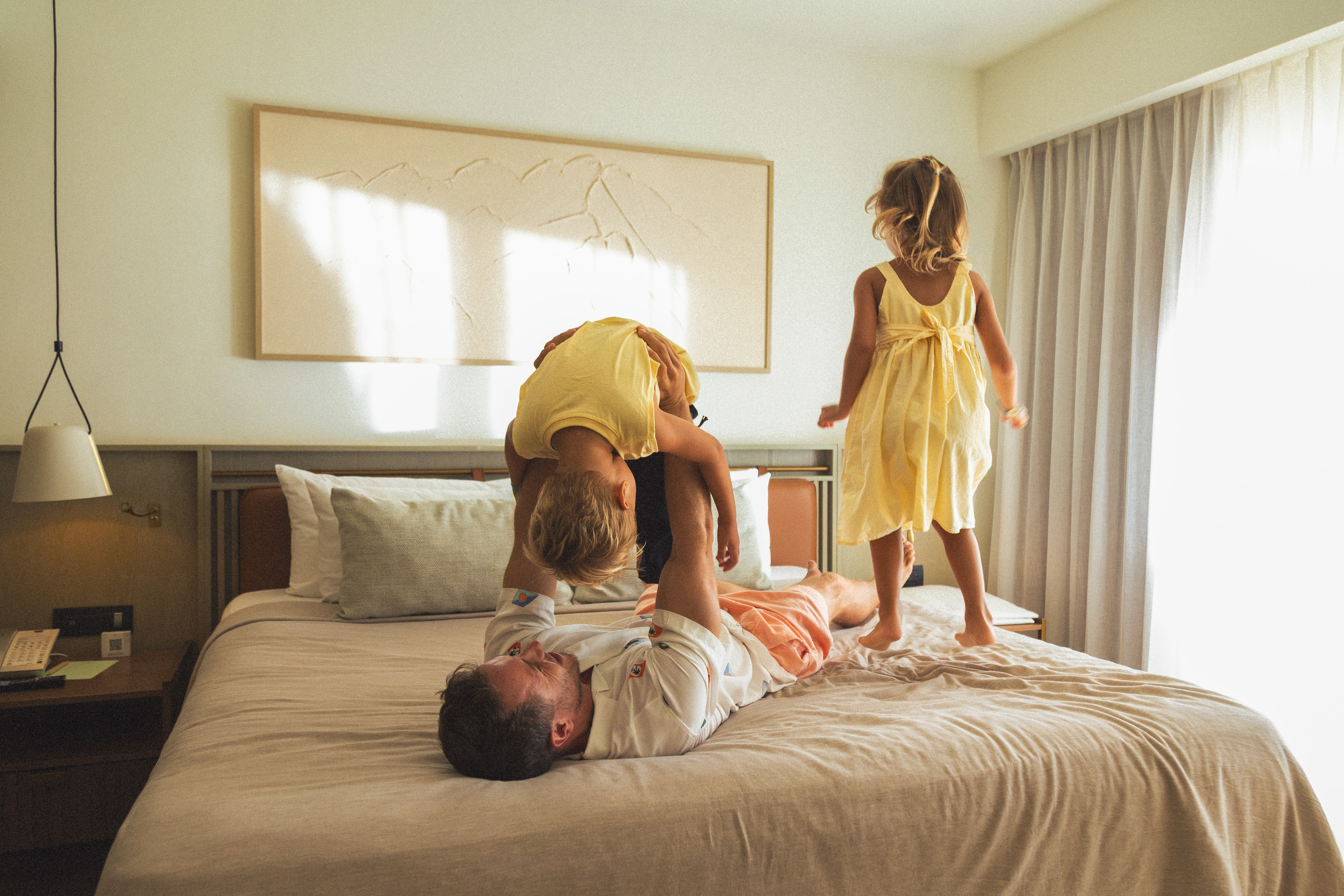 Playful dad with two kids on bed; one jumping, one held upside down.