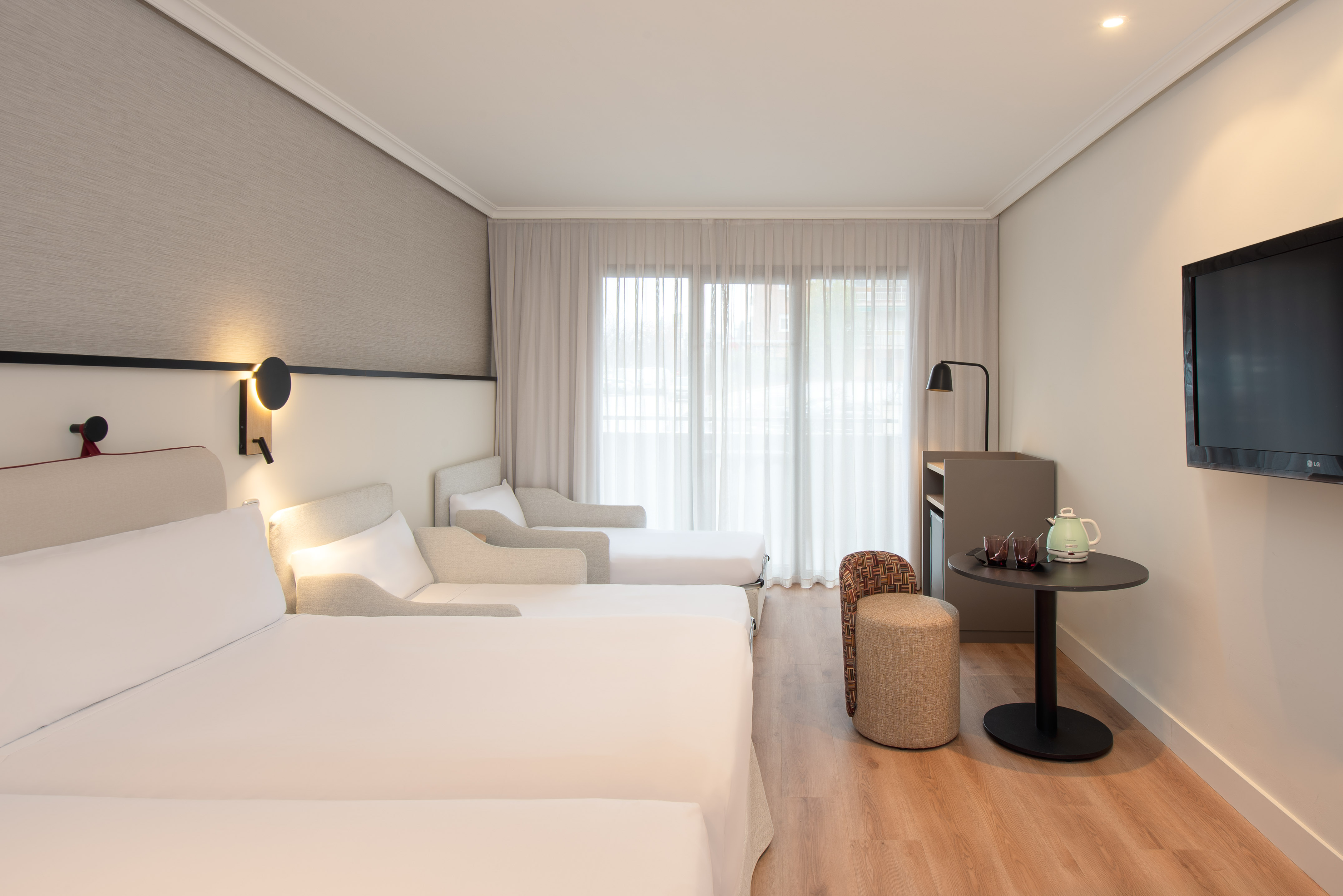 Modern hotel triple room with white beds, wooden floor, large window, and TV.