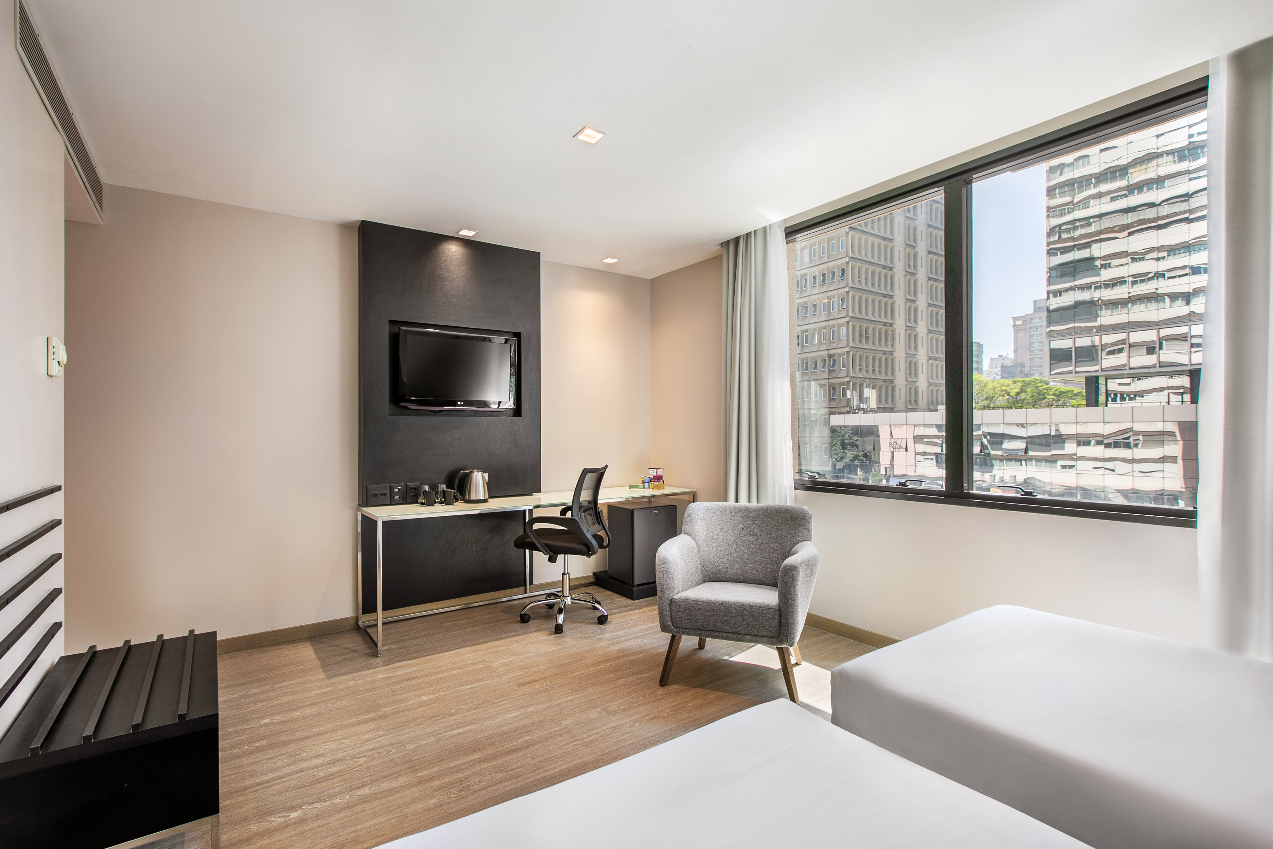 Modern hotel room with twin beds, city view, desk, TV, and armchair.