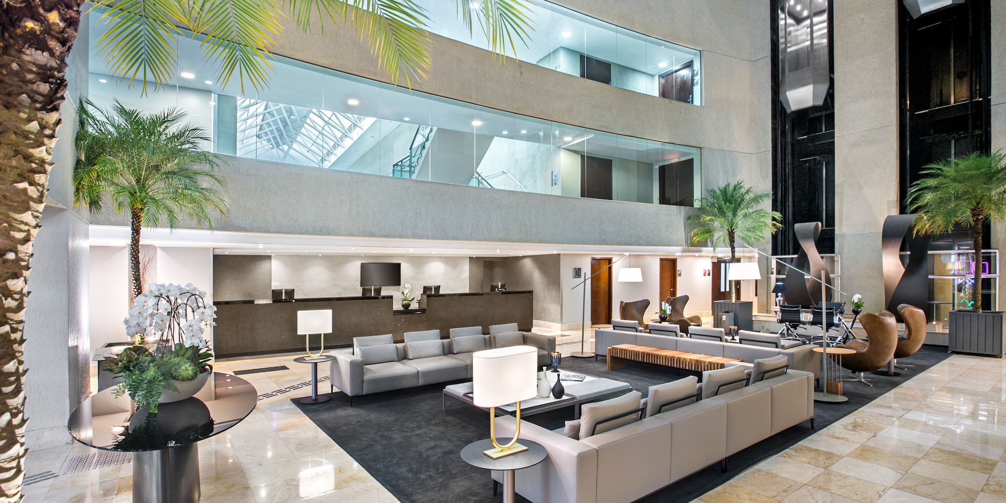 Modern hotel lobby lounge with reception, indoor palm trees, and glass elevators.