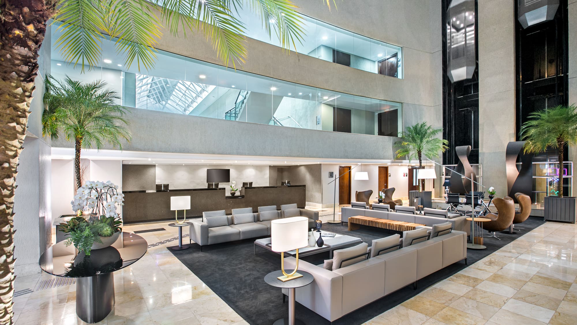 Modern hotel lobby with reception, glass elevators, and palm trees.