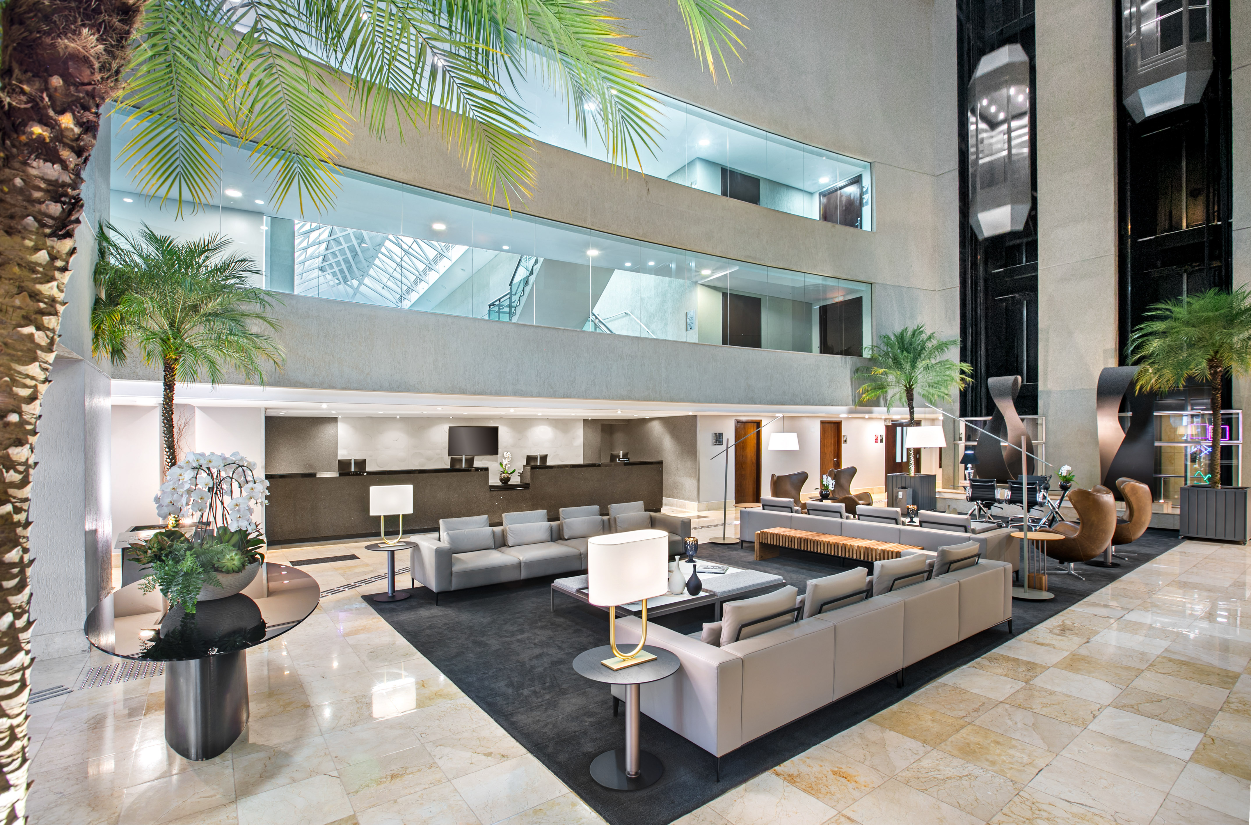 Modern hotel lobby lounge with reception, indoor palm trees, and glass elevators.