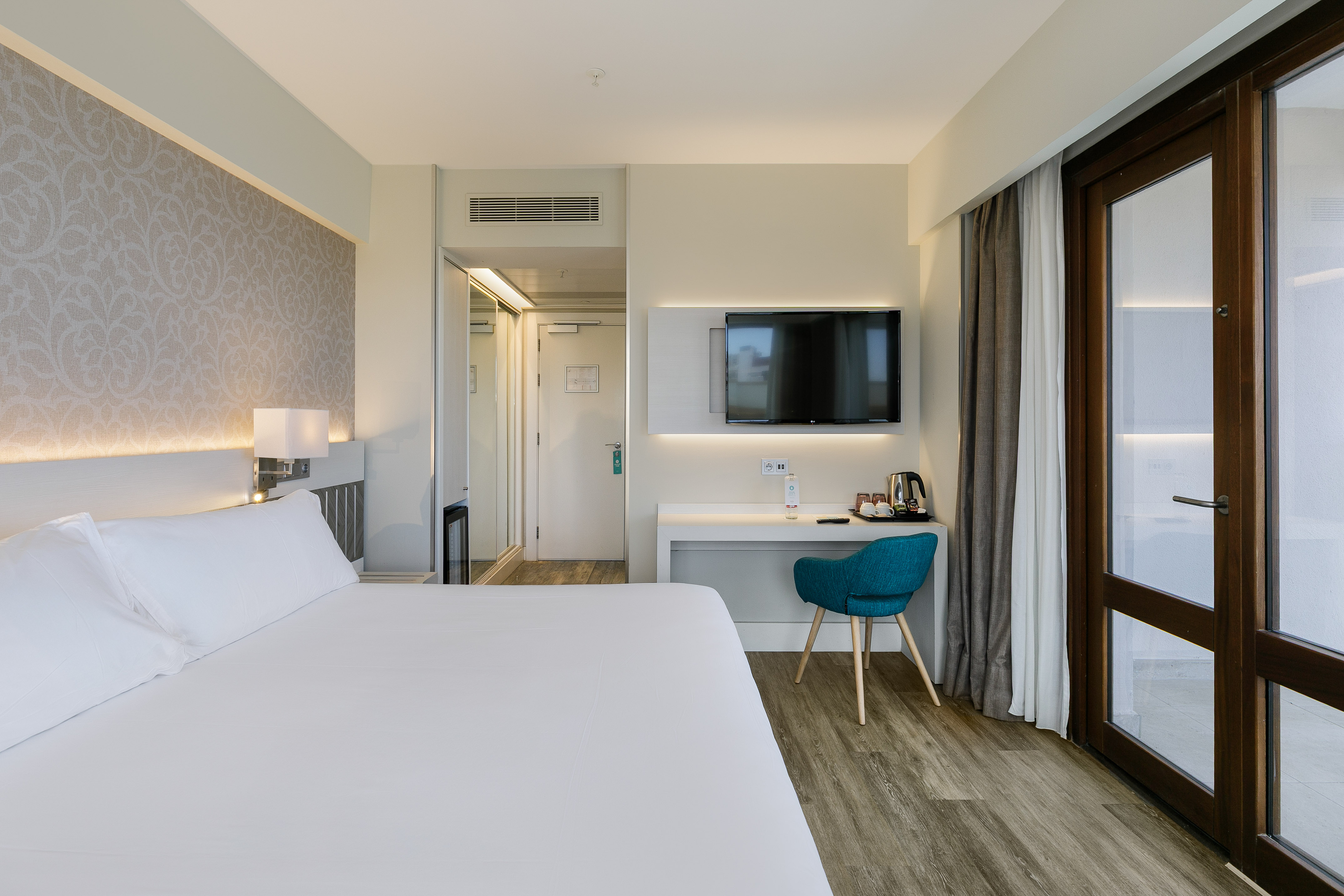 Modern hotel room with white bed, desk, wall TV, wood floor, and balcony doors.