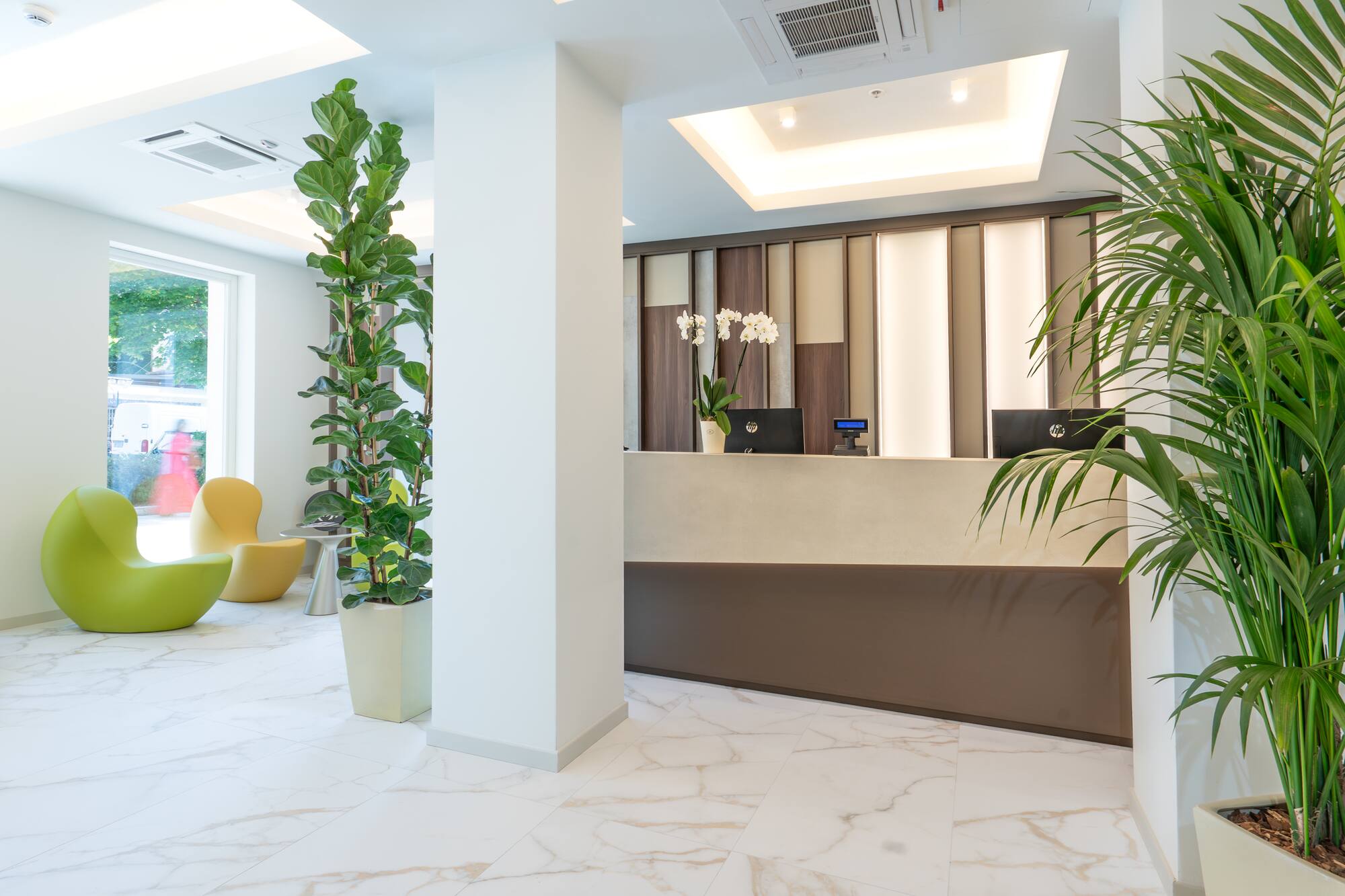 Bright hotel lobby with modern reception desk, white marble floor, and green plants.