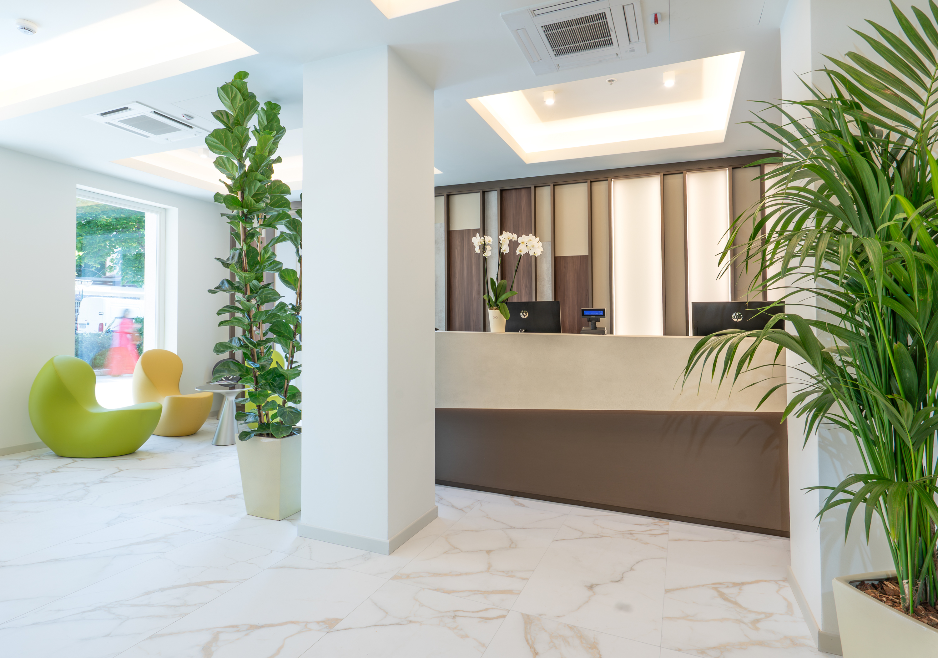 Bright hotel lobby with modern reception desk, white marble floor, and green plants.
