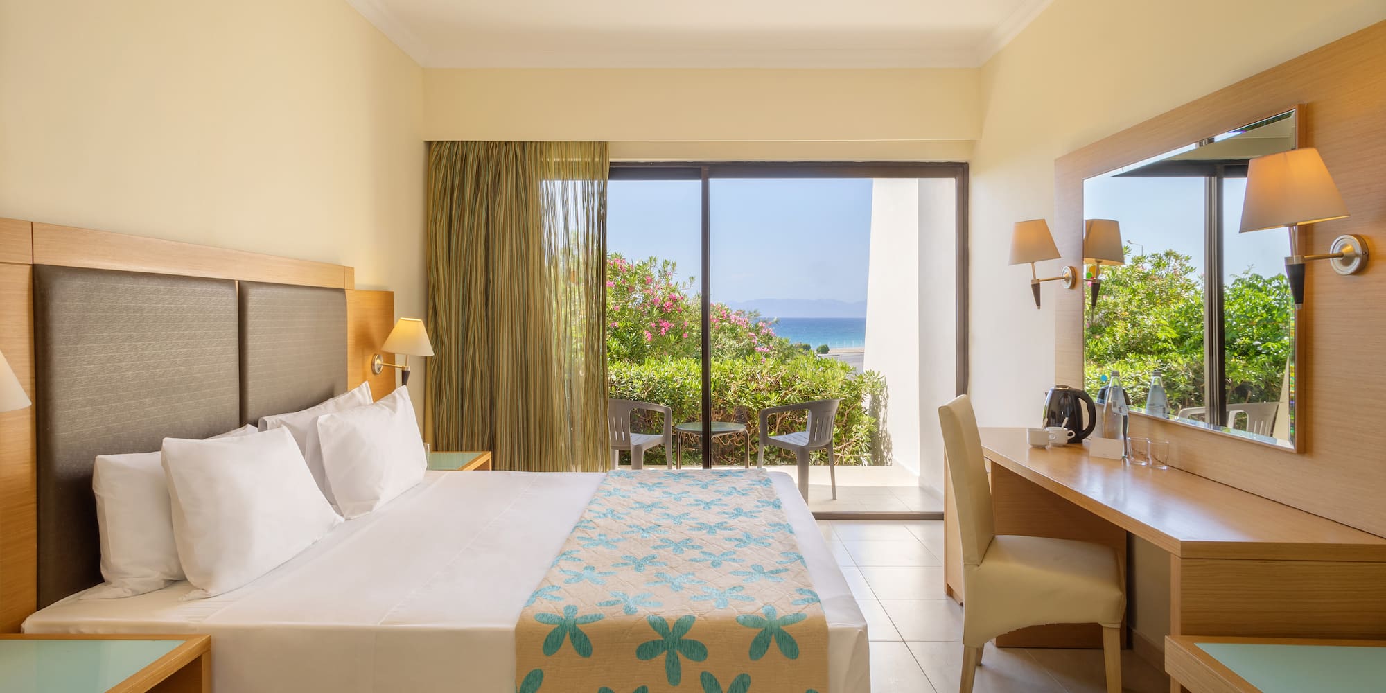 Modern hotel room with bed, desk, and private balcony overlooking the ocean.