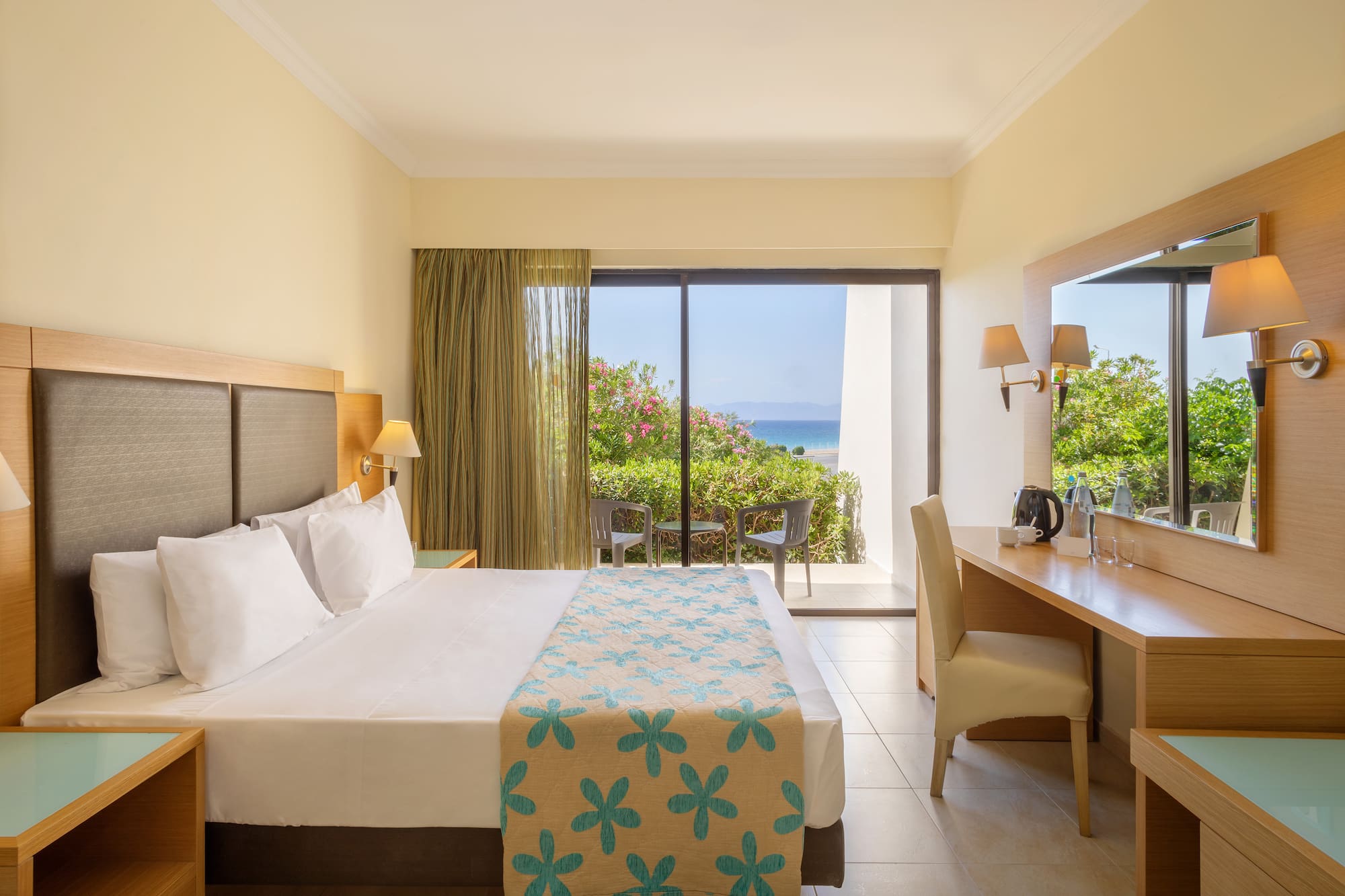 Modern hotel room with bed, desk, and private balcony overlooking the ocean.