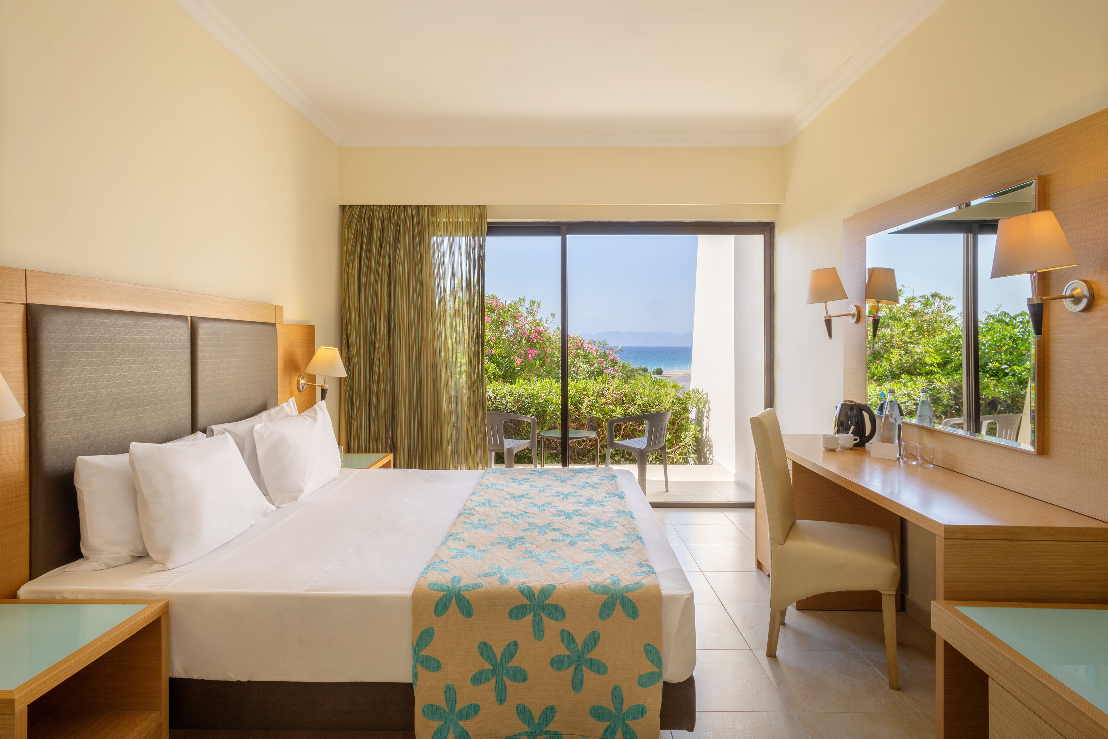Modern hotel room with bed, desk, and private balcony overlooking the ocean.