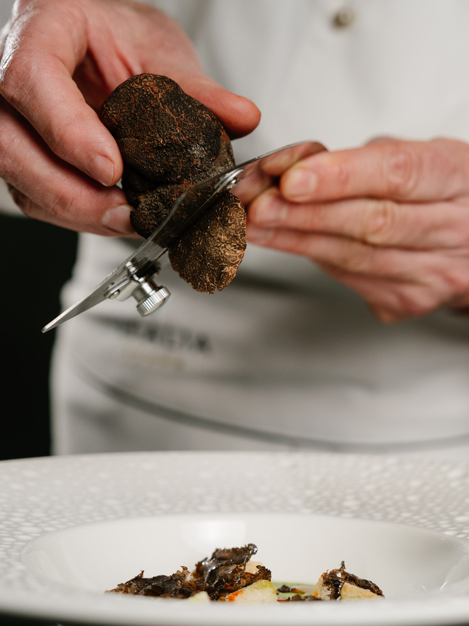 Chef hands shaving black truffle with a slicer onto a white gourmet plate.