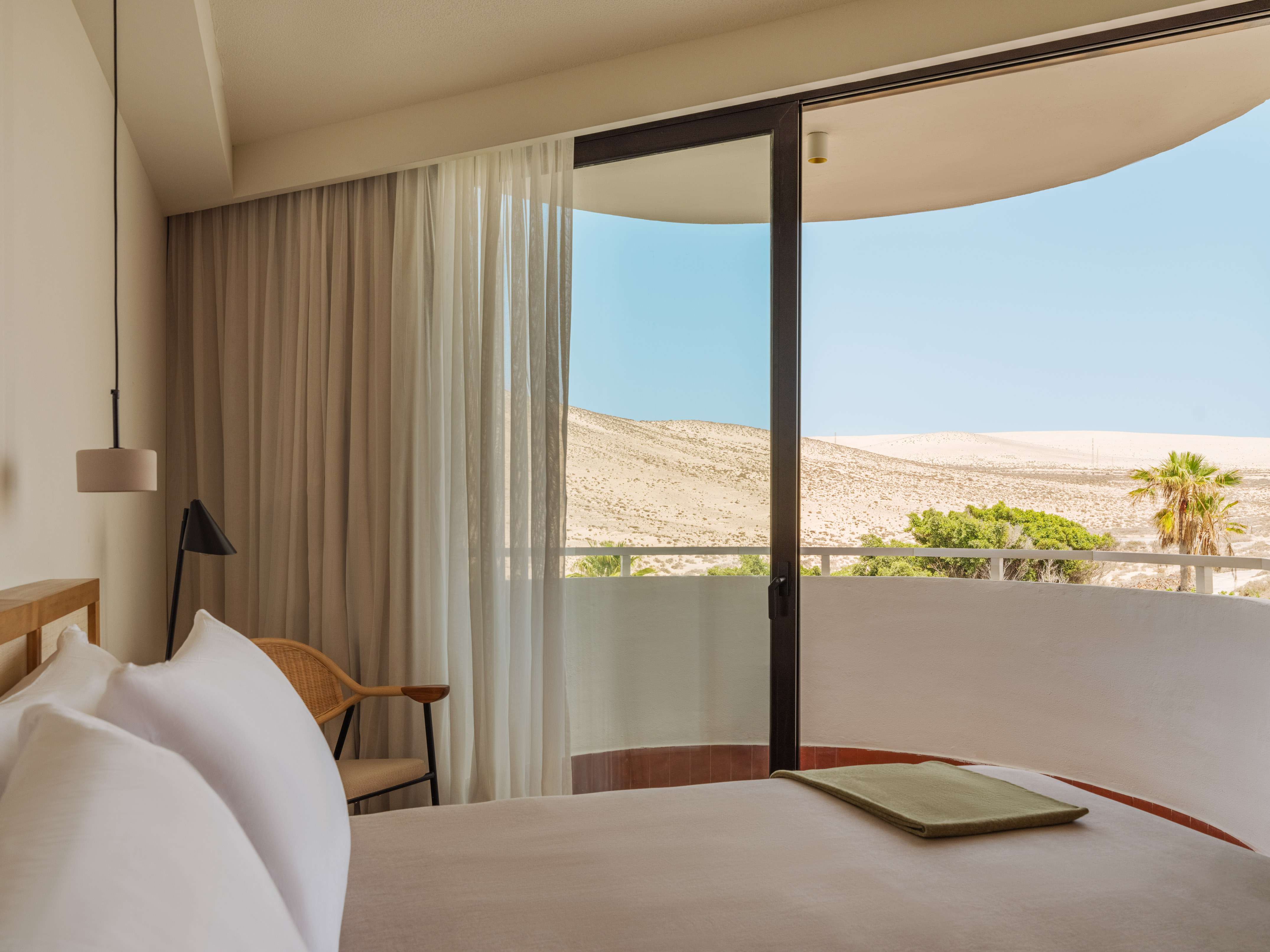 Hotel bedroom with balcony overlooking desert dunes and palm trees.