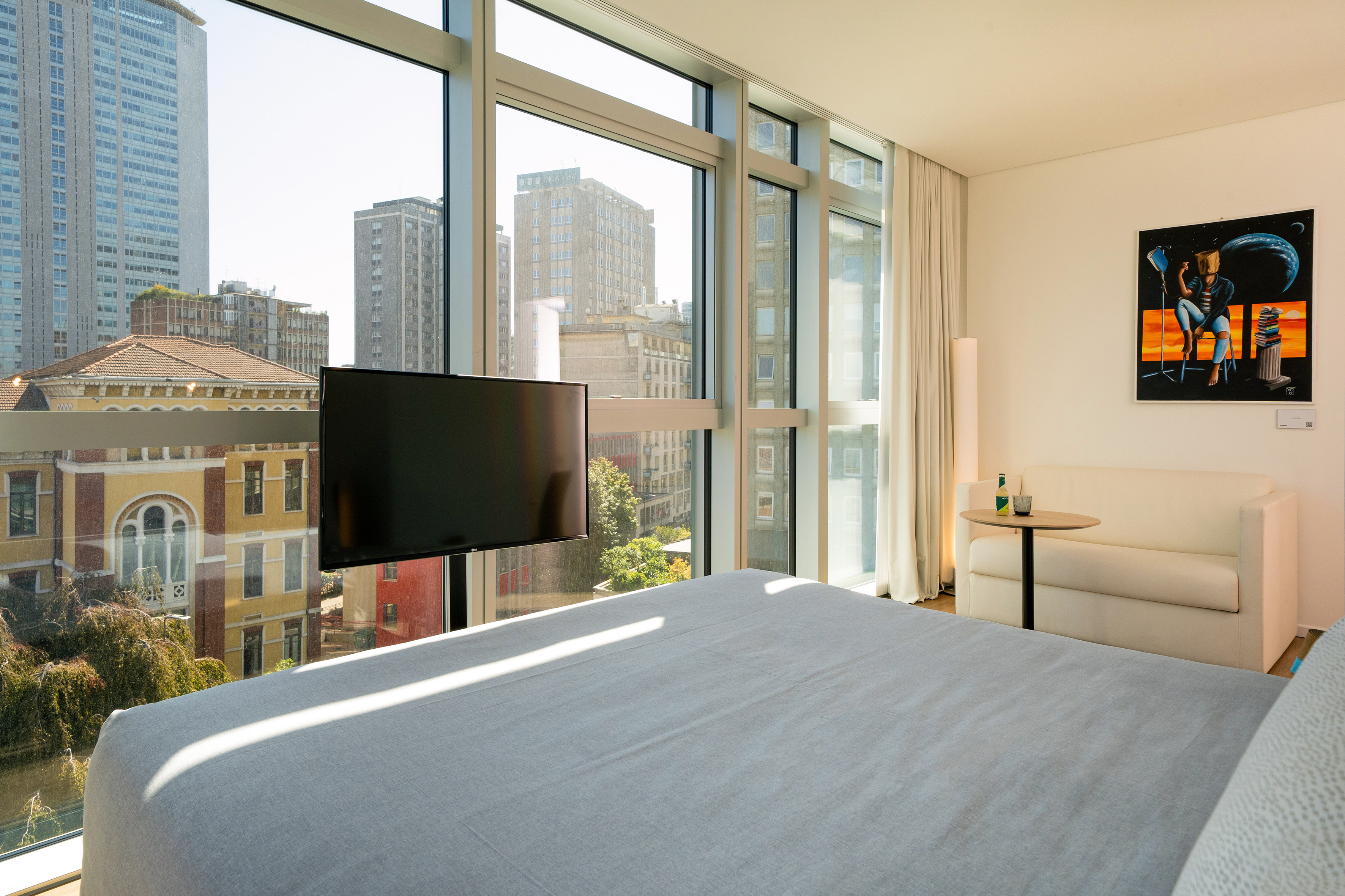 Modern hotel room with city view, large window, bed, TV, and sofa.