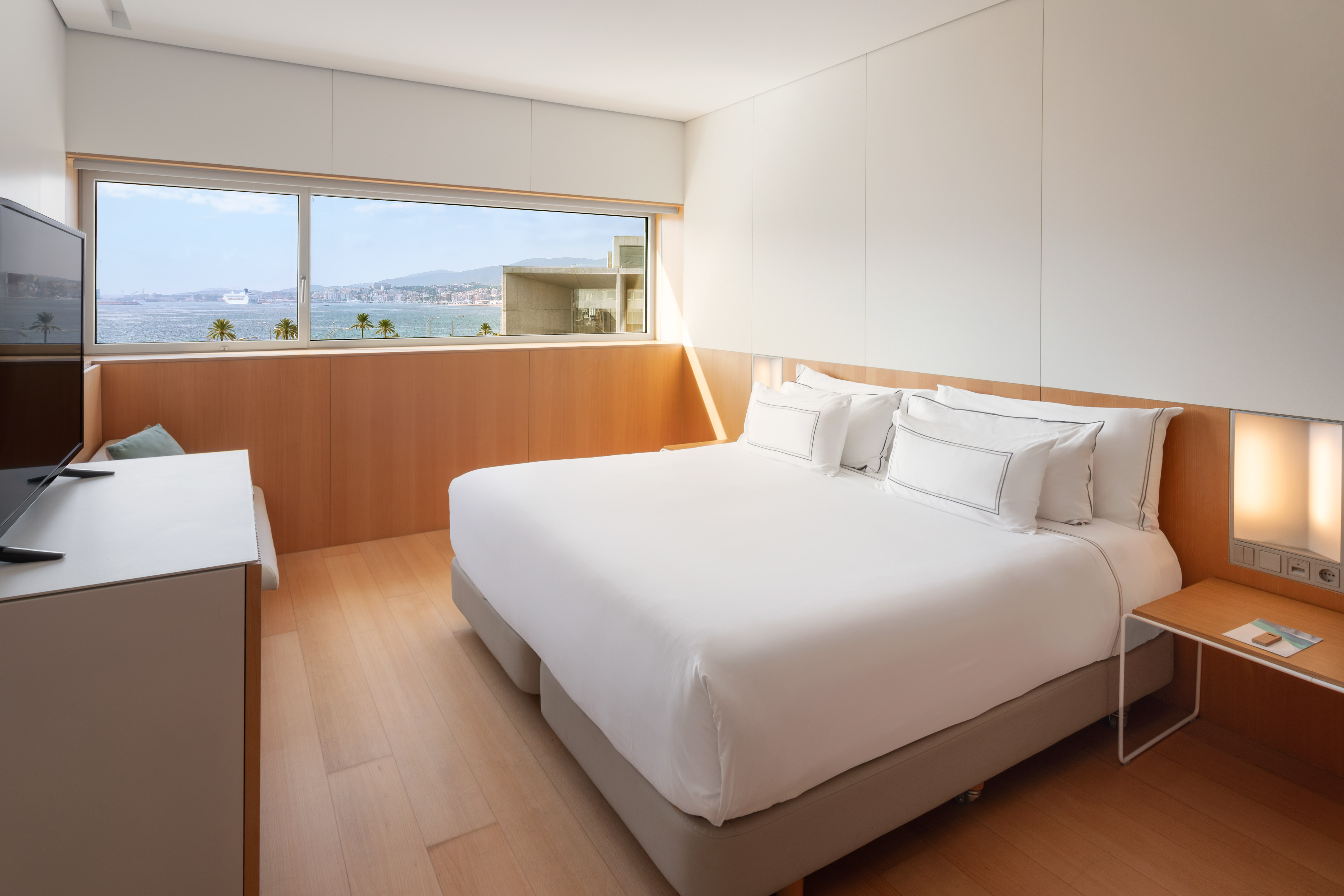Modern hotel room with panoramic sea view, white bed, and wooden floor.