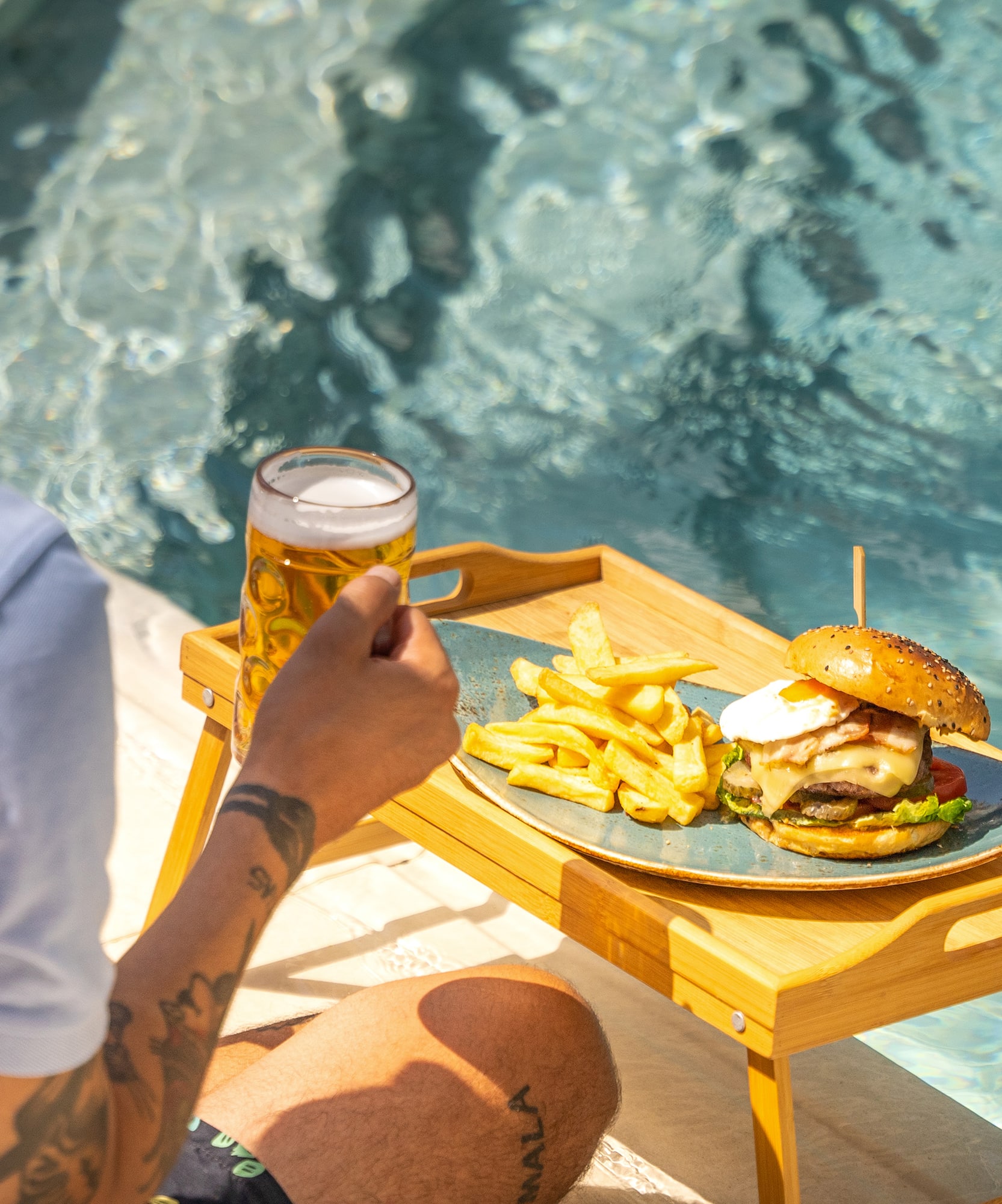 a person sitting by a pool holding a glass of beer and a burger and fries