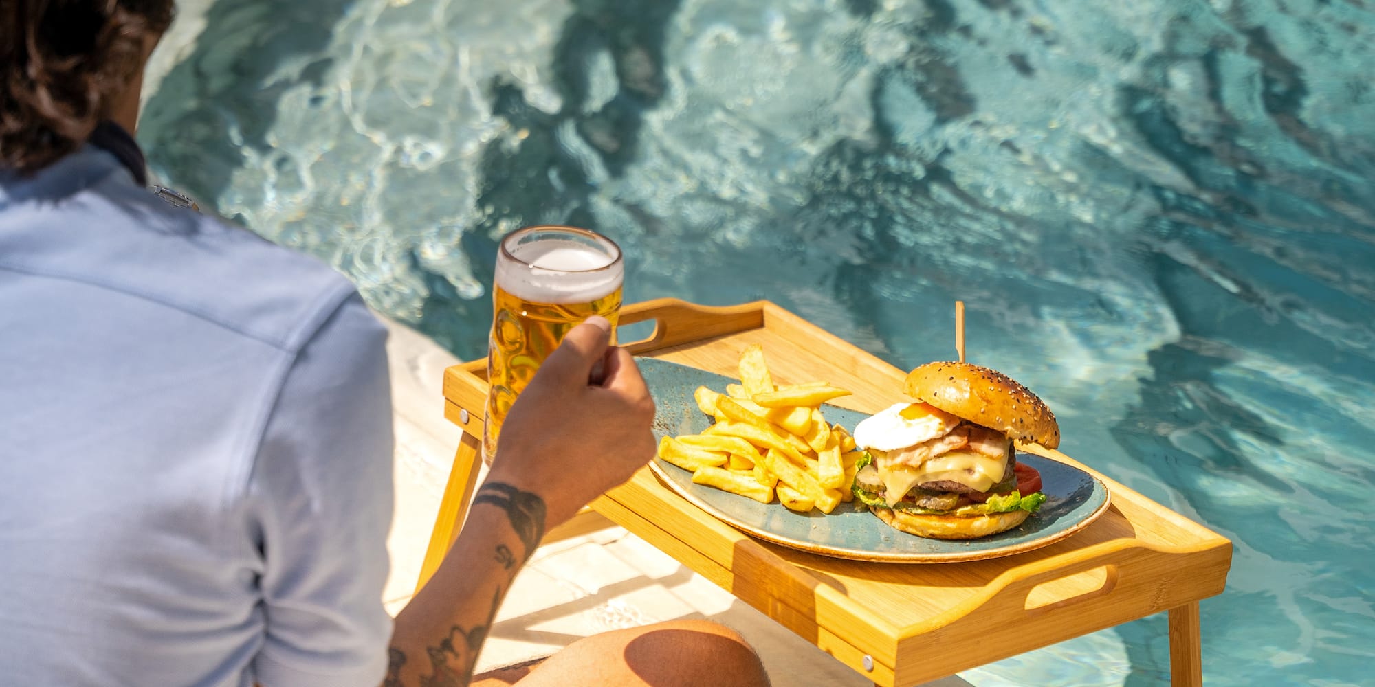 a person sitting by a pool holding a glass of beer and a burger and fries