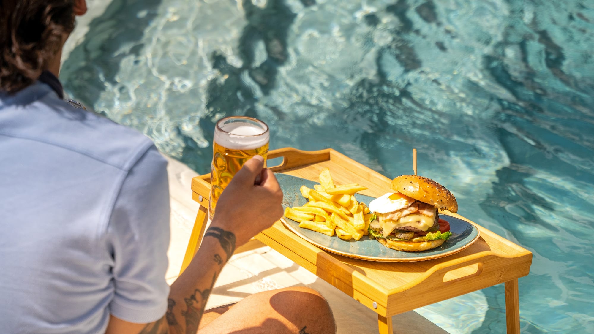 a person sitting by a pool holding a glass of beer and a burger and fries
