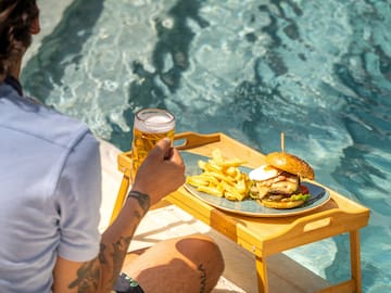 a person sitting by a pool holding a glass of beer and a burger and fries