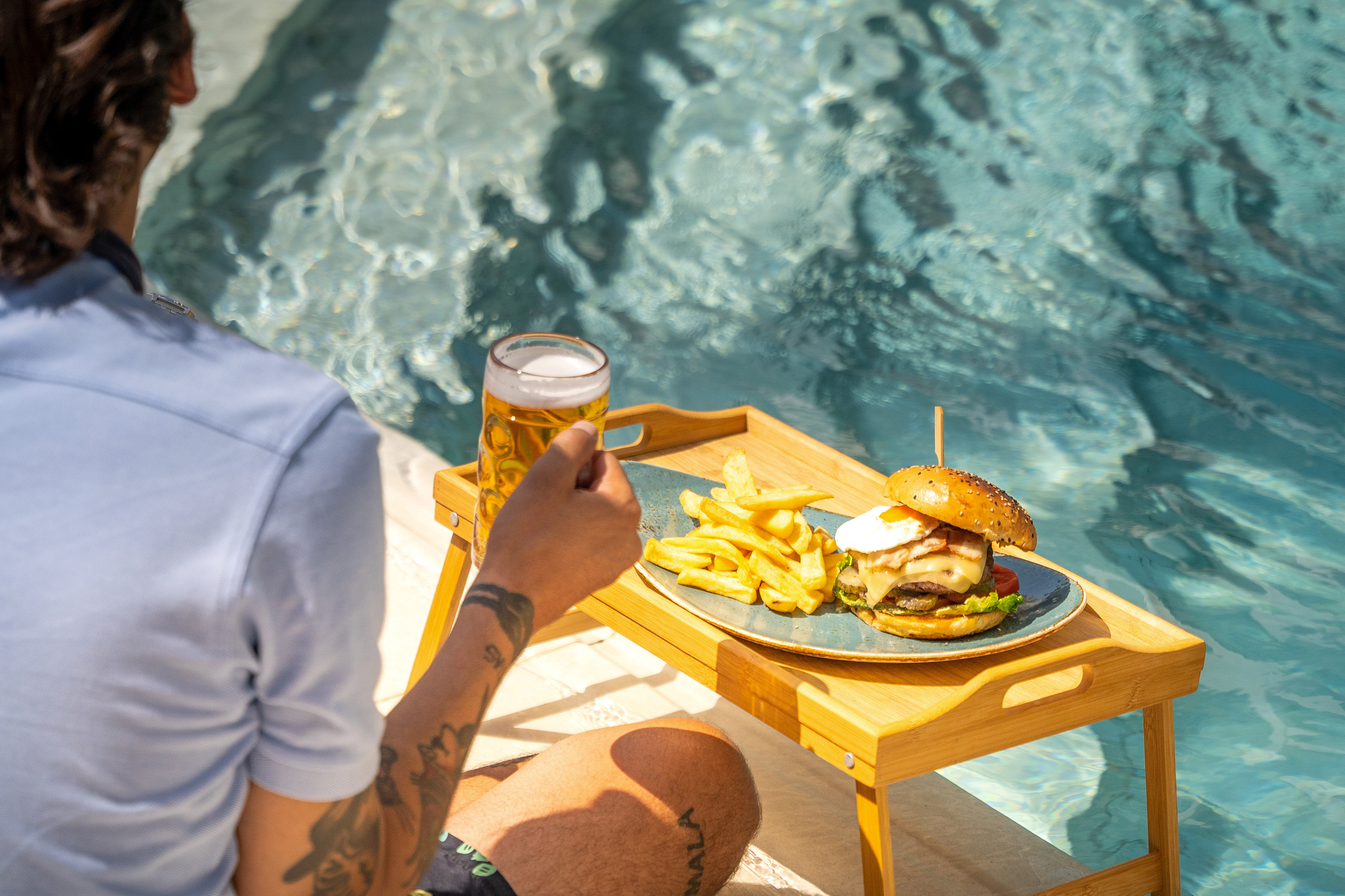 Luxury poolside dining: burger, fries, and beer on a wooden tray.