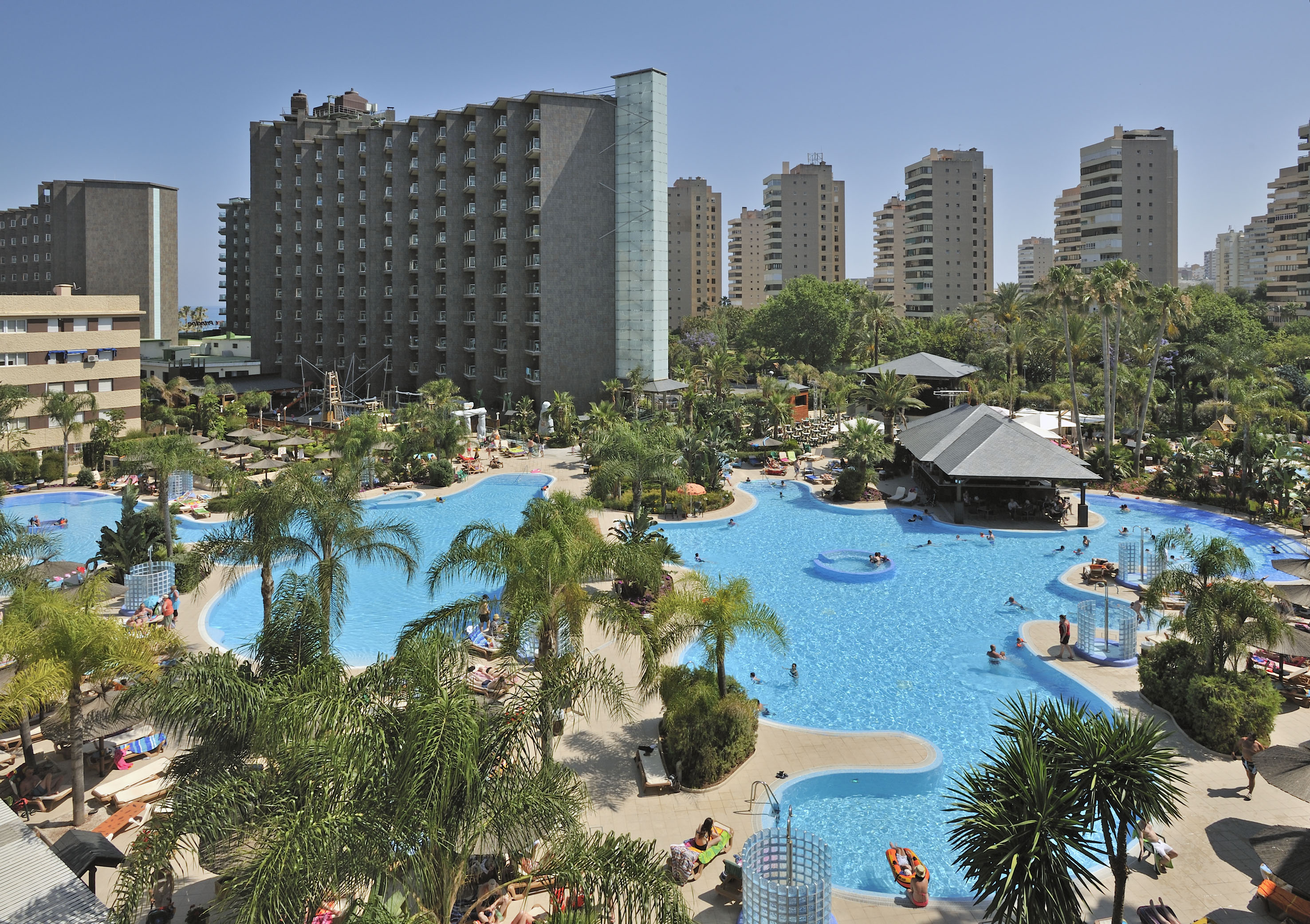 Resort con grandes piscinas, palmeras y edificios hoteleros bajo el sol.
