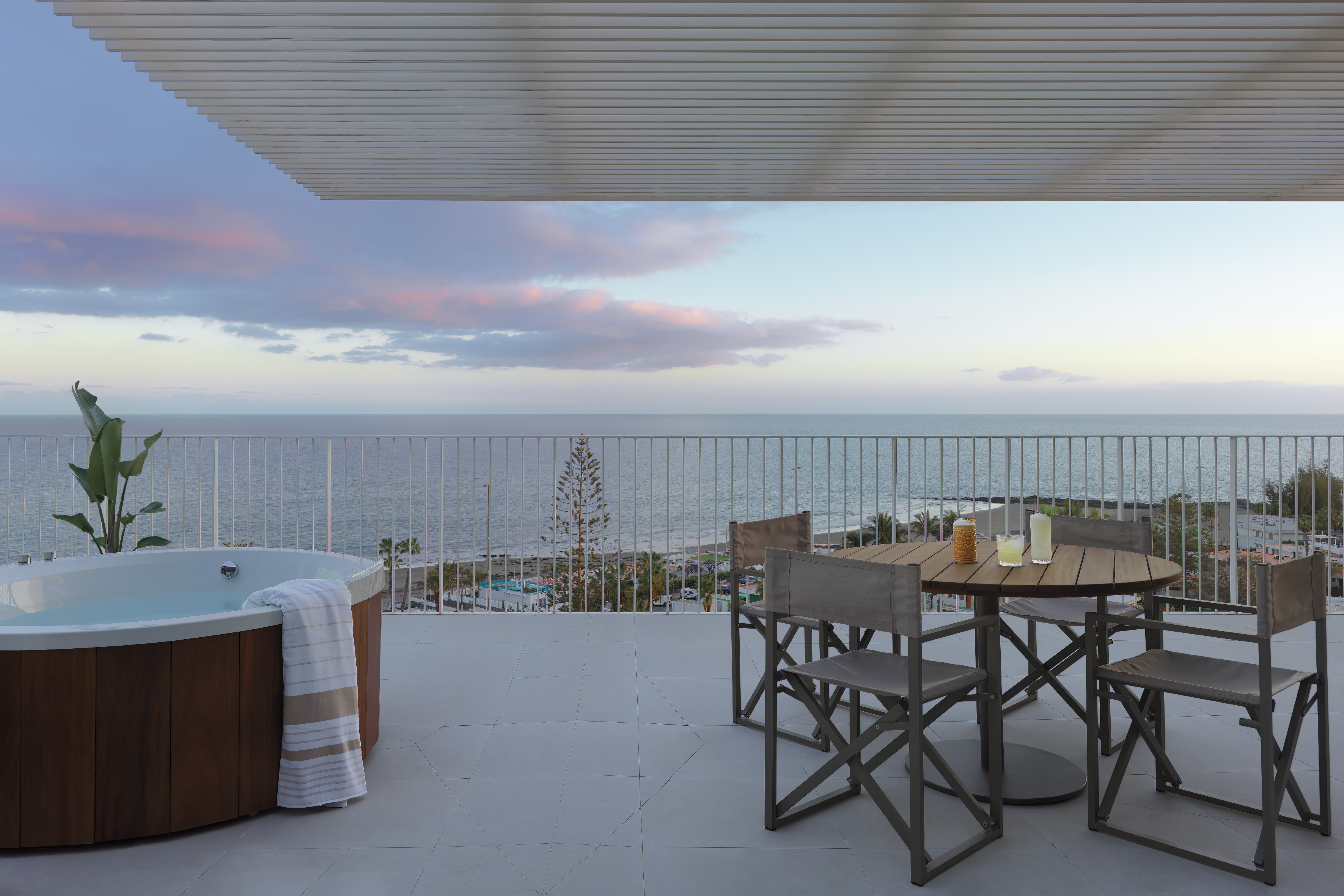 Balcony with hot tub, dining area, and ocean view at sunset.