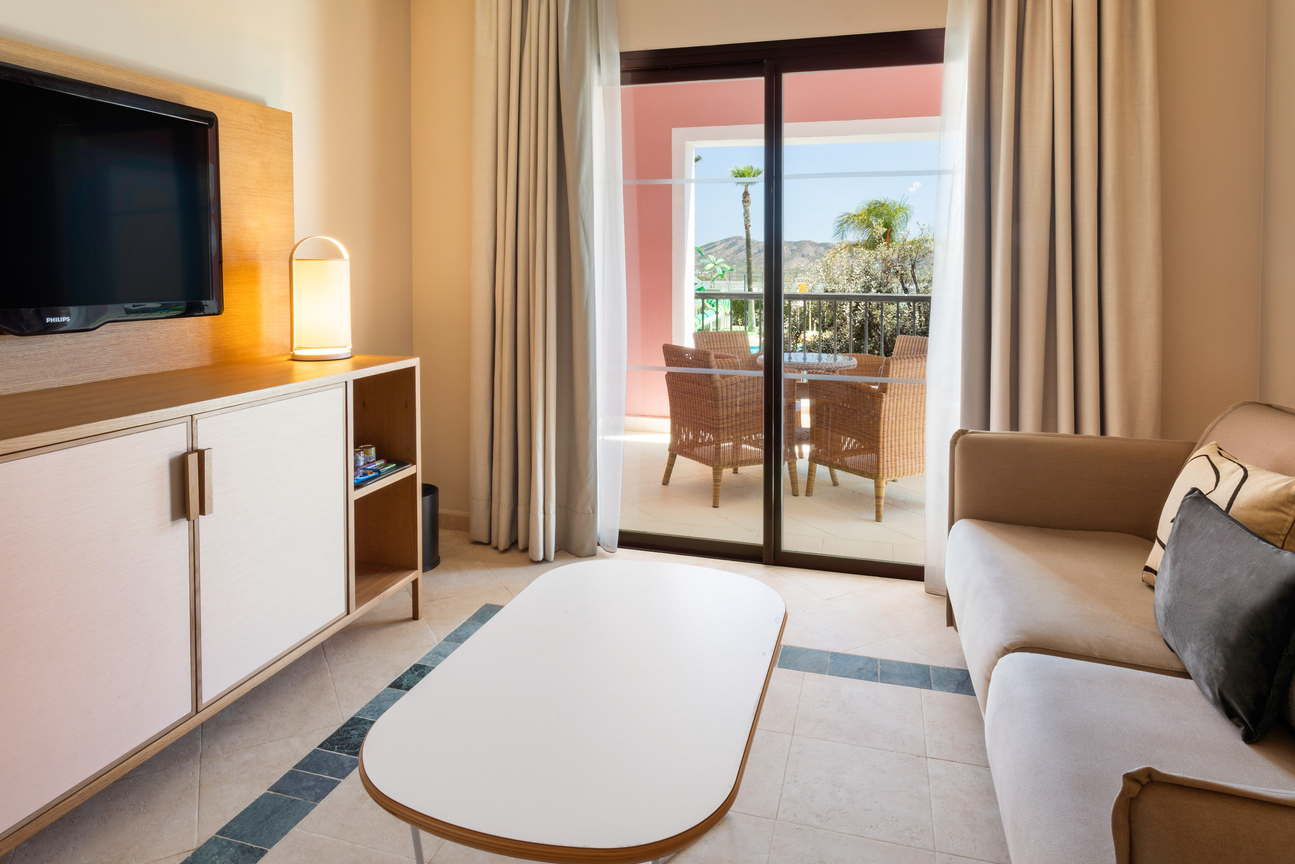 Bright hotel living room with balcony, wicker furniture, and mountain view.
