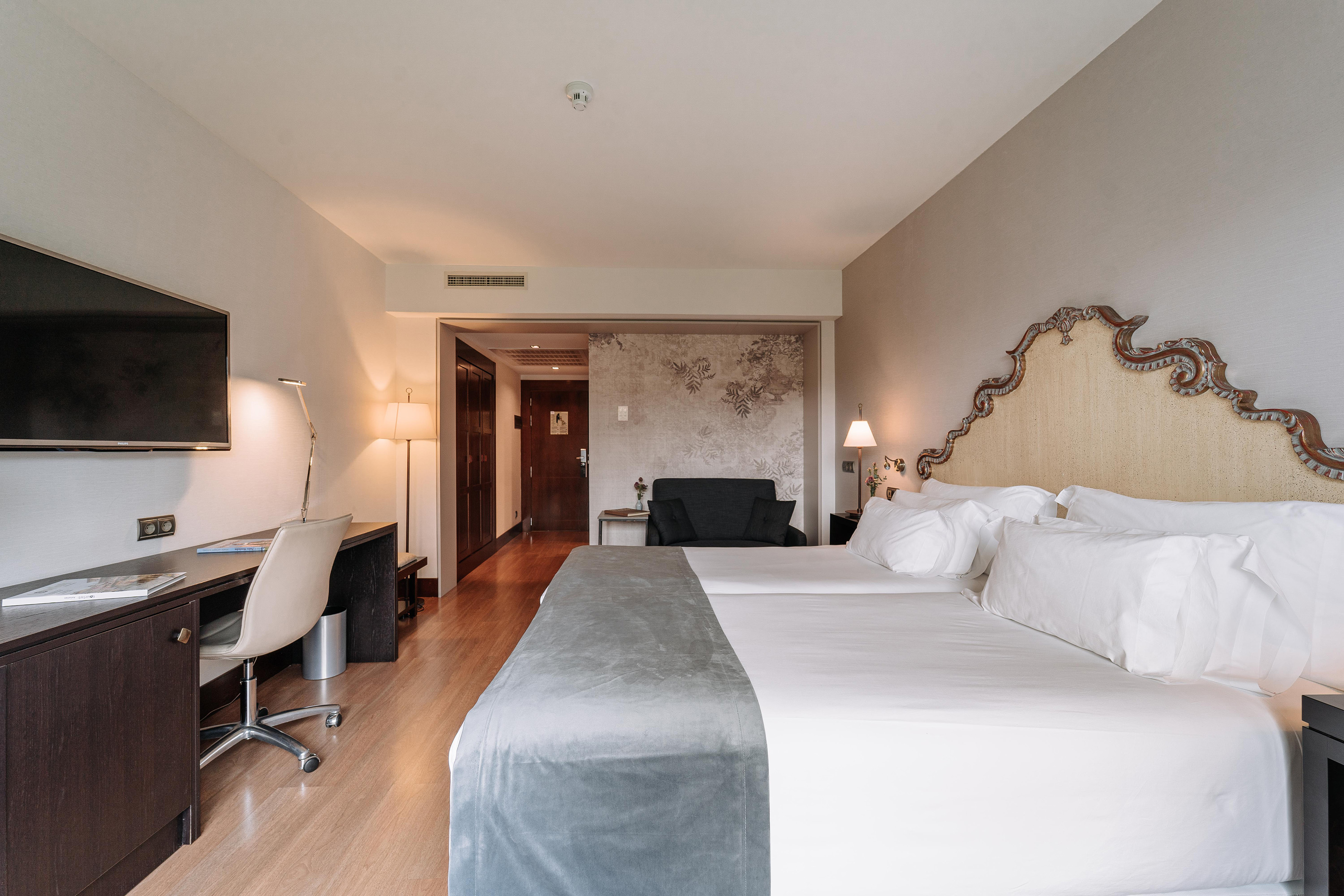 Hotel room with two beds, ornate headboard, desk, TV, and adjacent lounge area.
