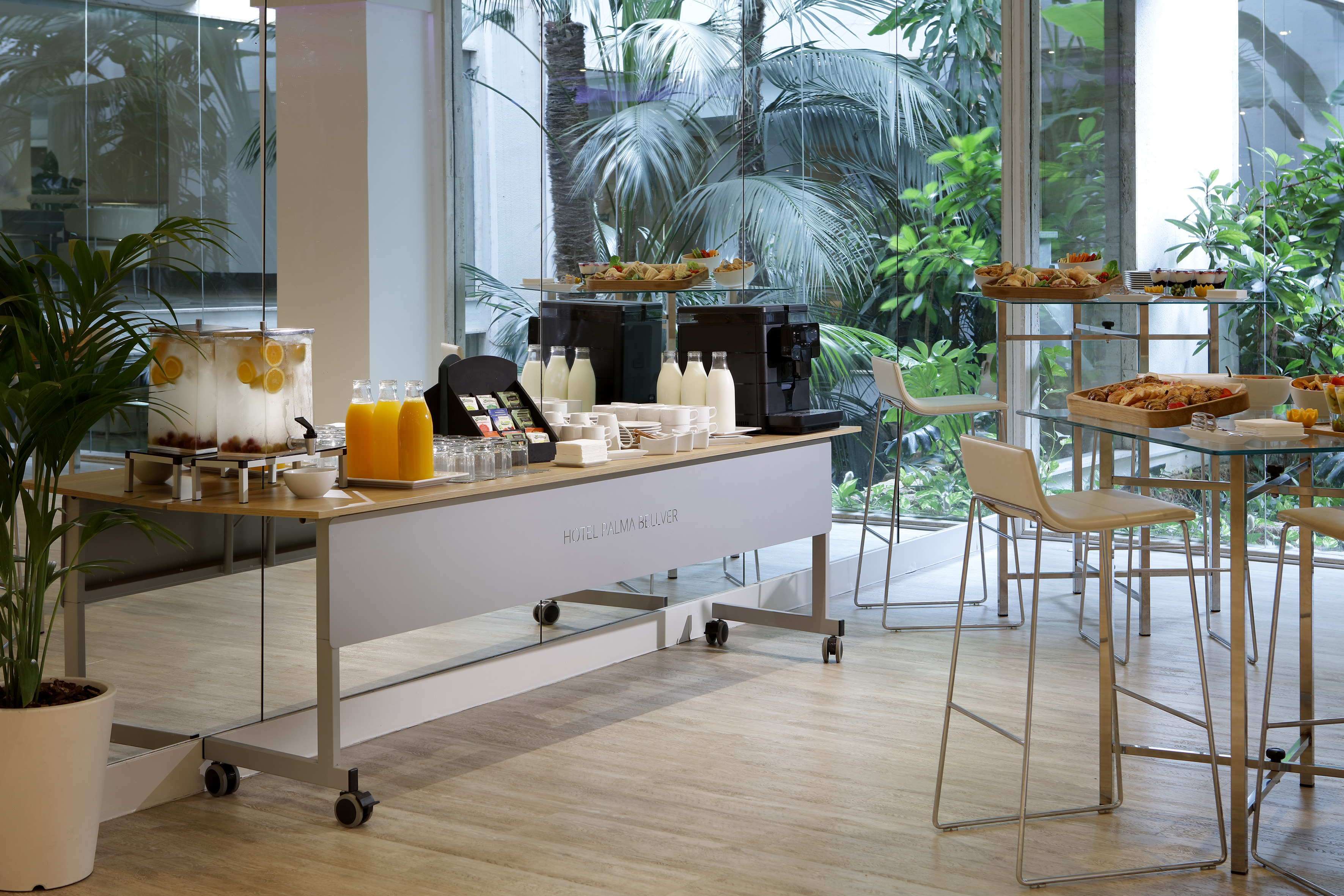 Modern hotel breakfast buffet with juices, coffee, food, and tropical greenery view.