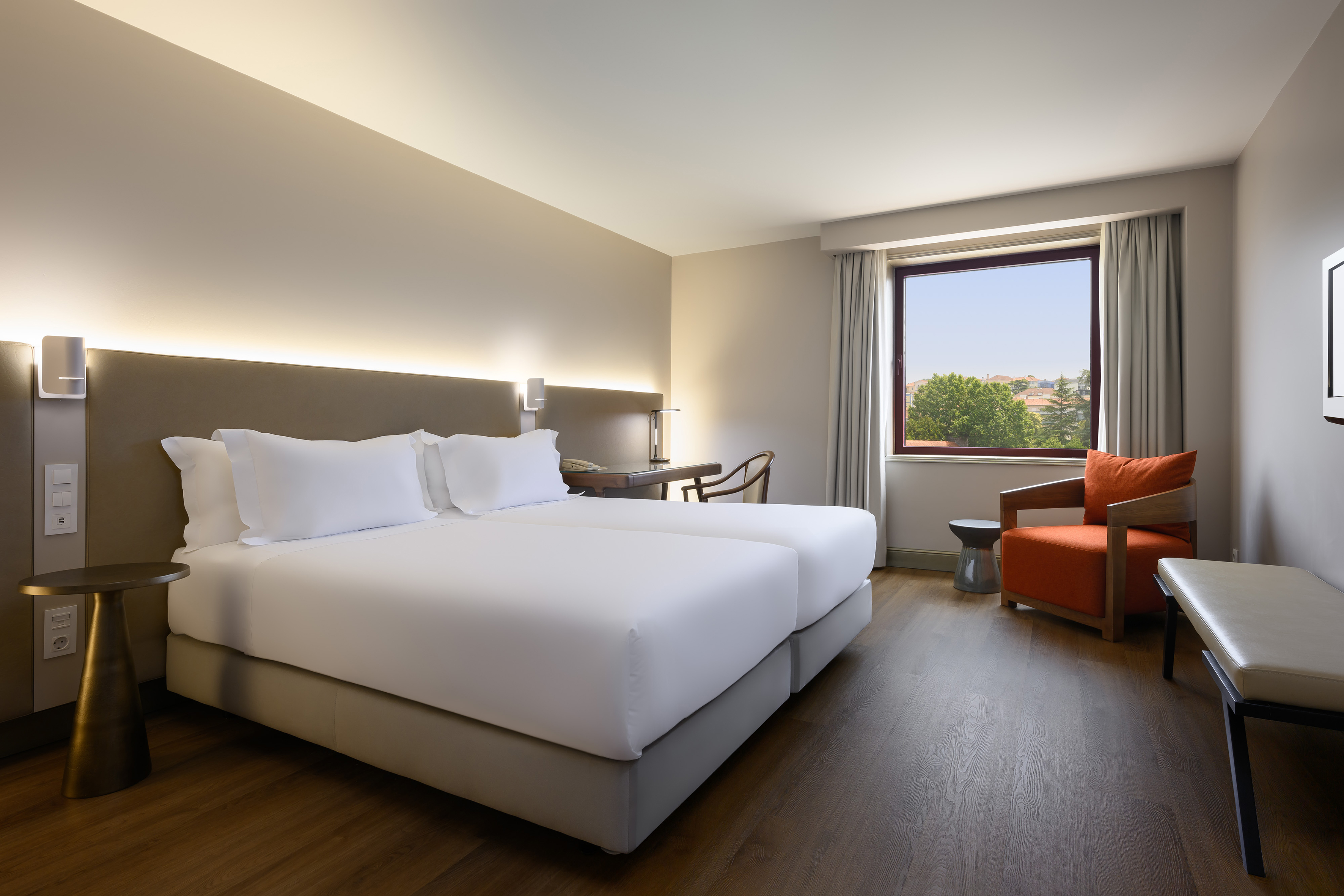 Modern hotel room with twin beds, desk, orange armchair, and window view.