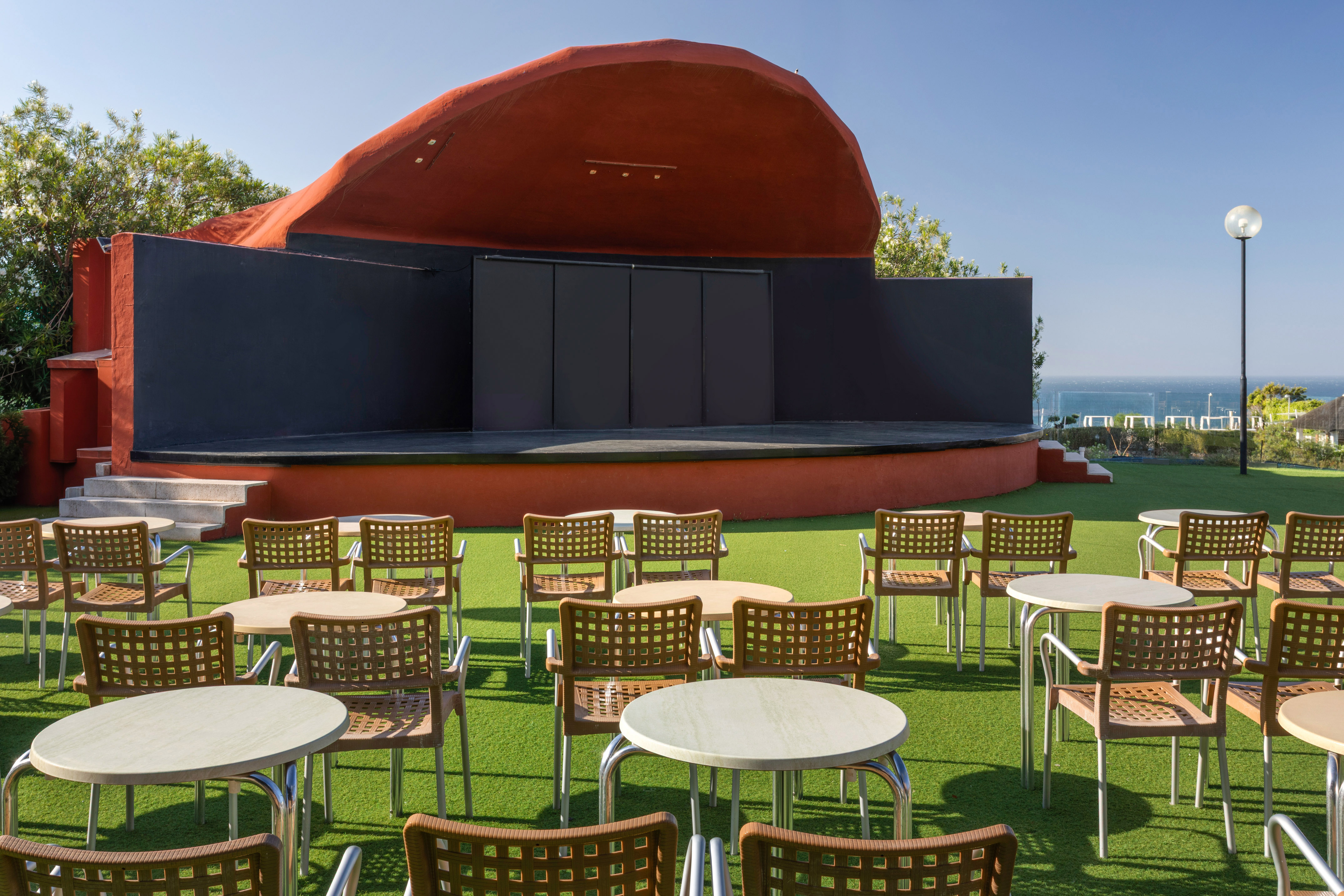 Outdoor amphitheater stage with tables, chairs on green turf, and ocean view.