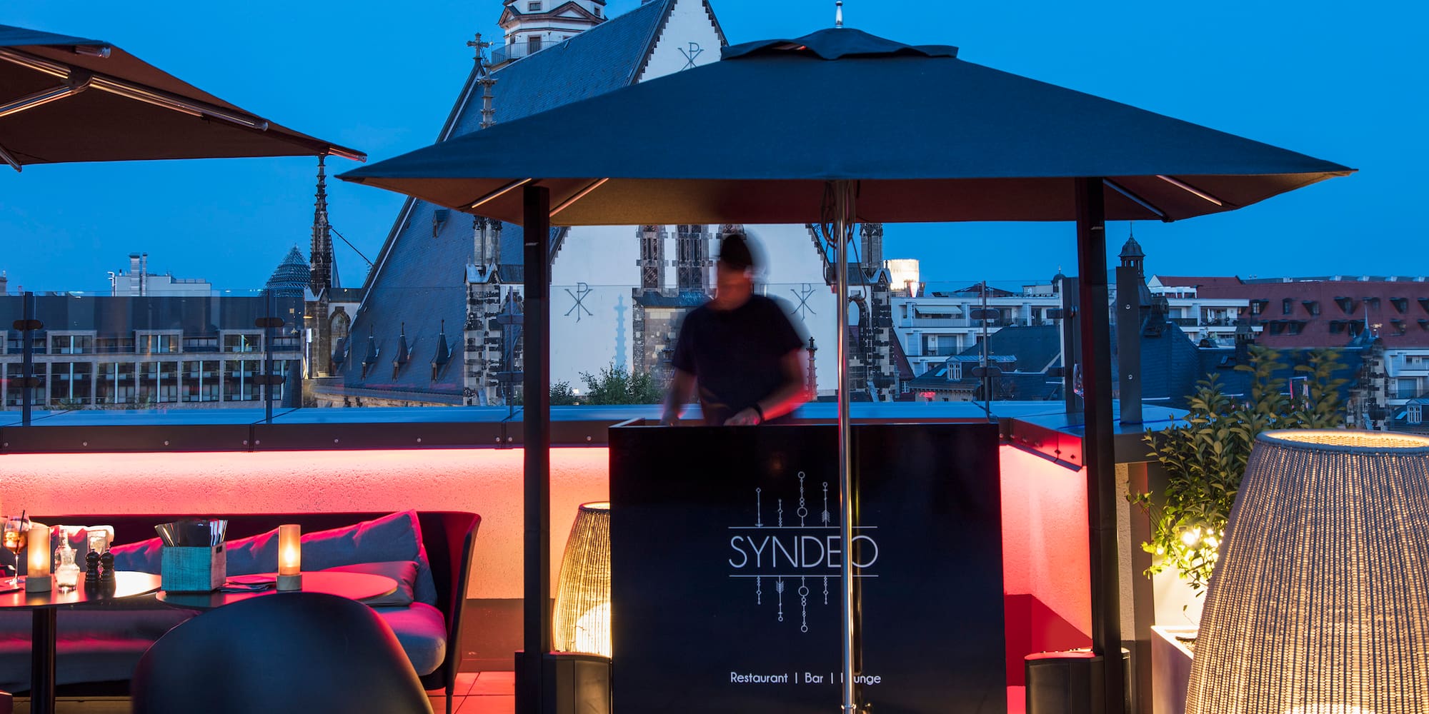 SYNDEO rooftop bar lounge with DJ, modern seating, and city view at dusk.