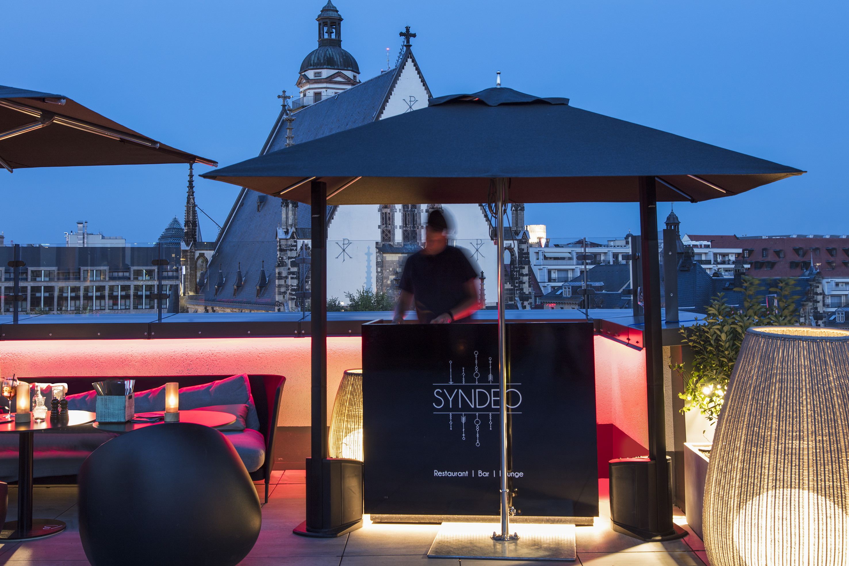 SYNDEO rooftop bar lounge with DJ, modern seating, and city view at dusk.