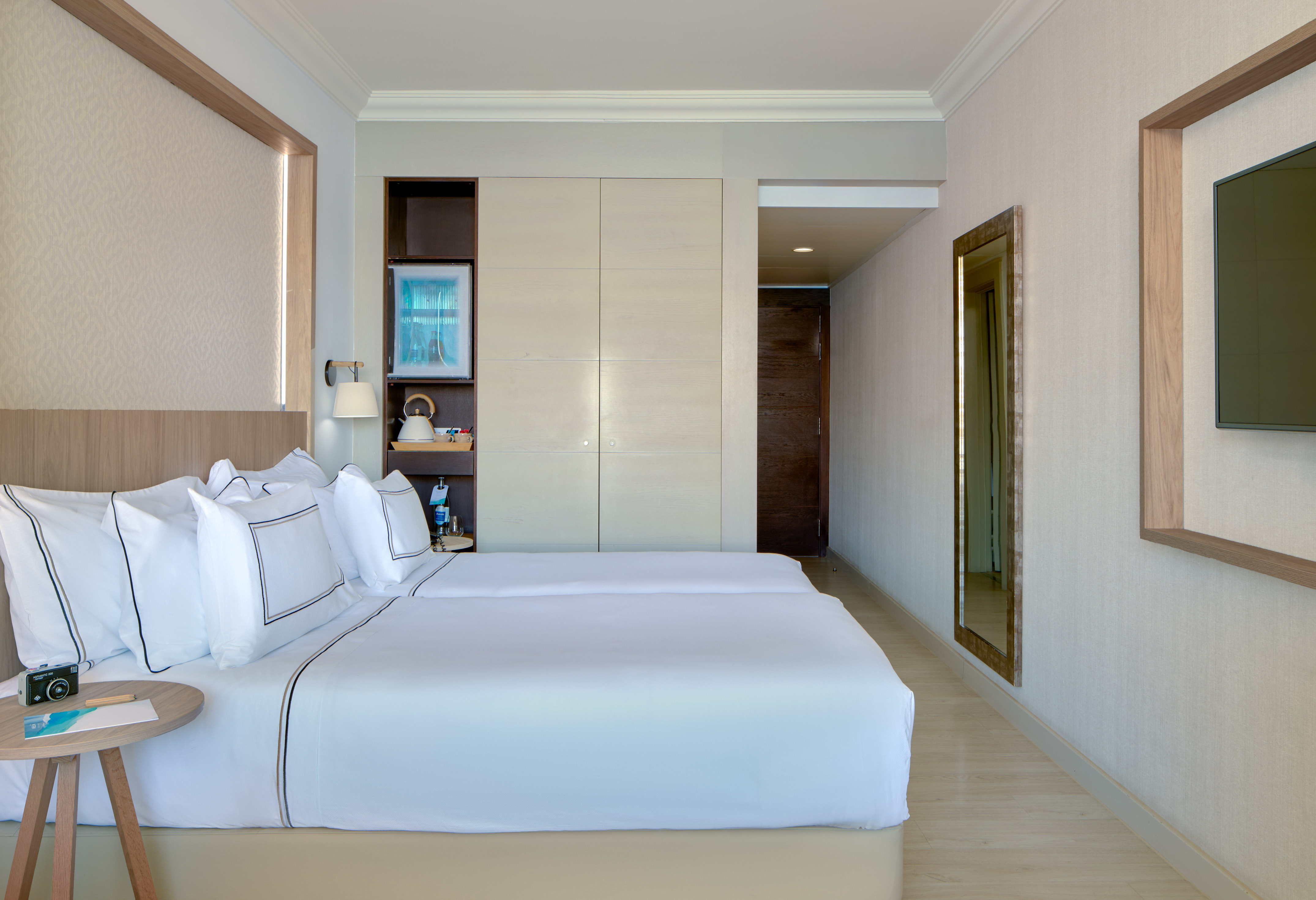 Modern hotel room with two twin beds, minibar, TV, and full-length mirror.