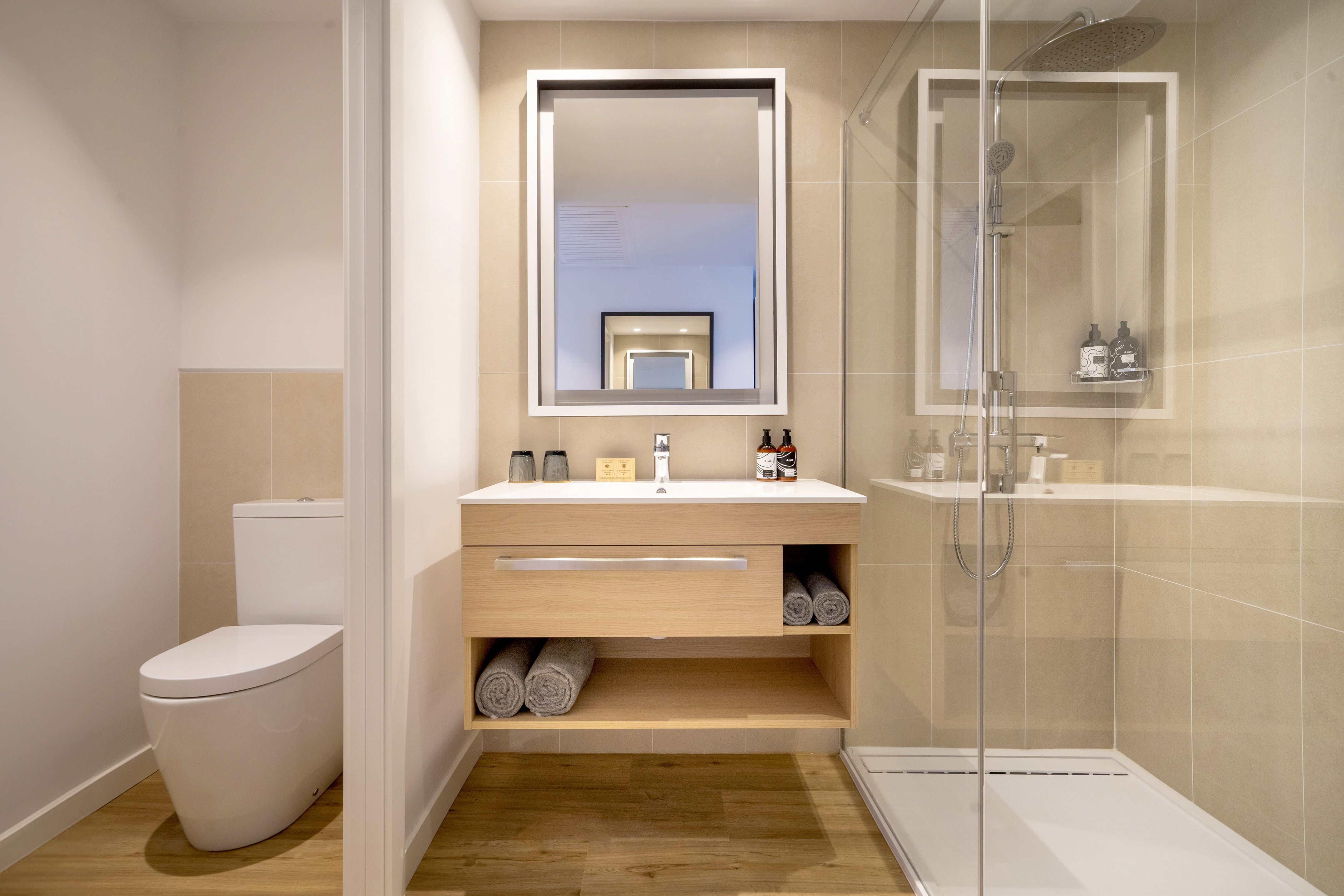 Modern bathroom with wooden vanity, sink, mirror, glass shower, and toilet area.