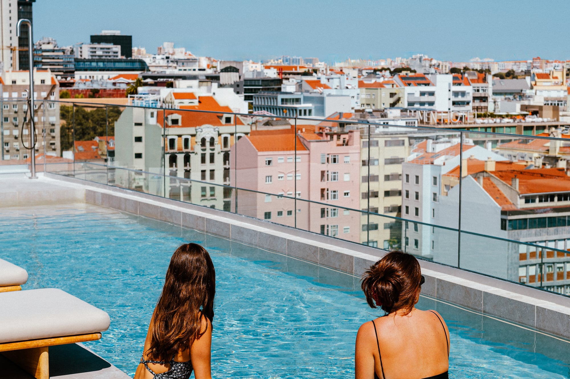 Women relaxing at a sunny rooftop pool with a panoramic city view.