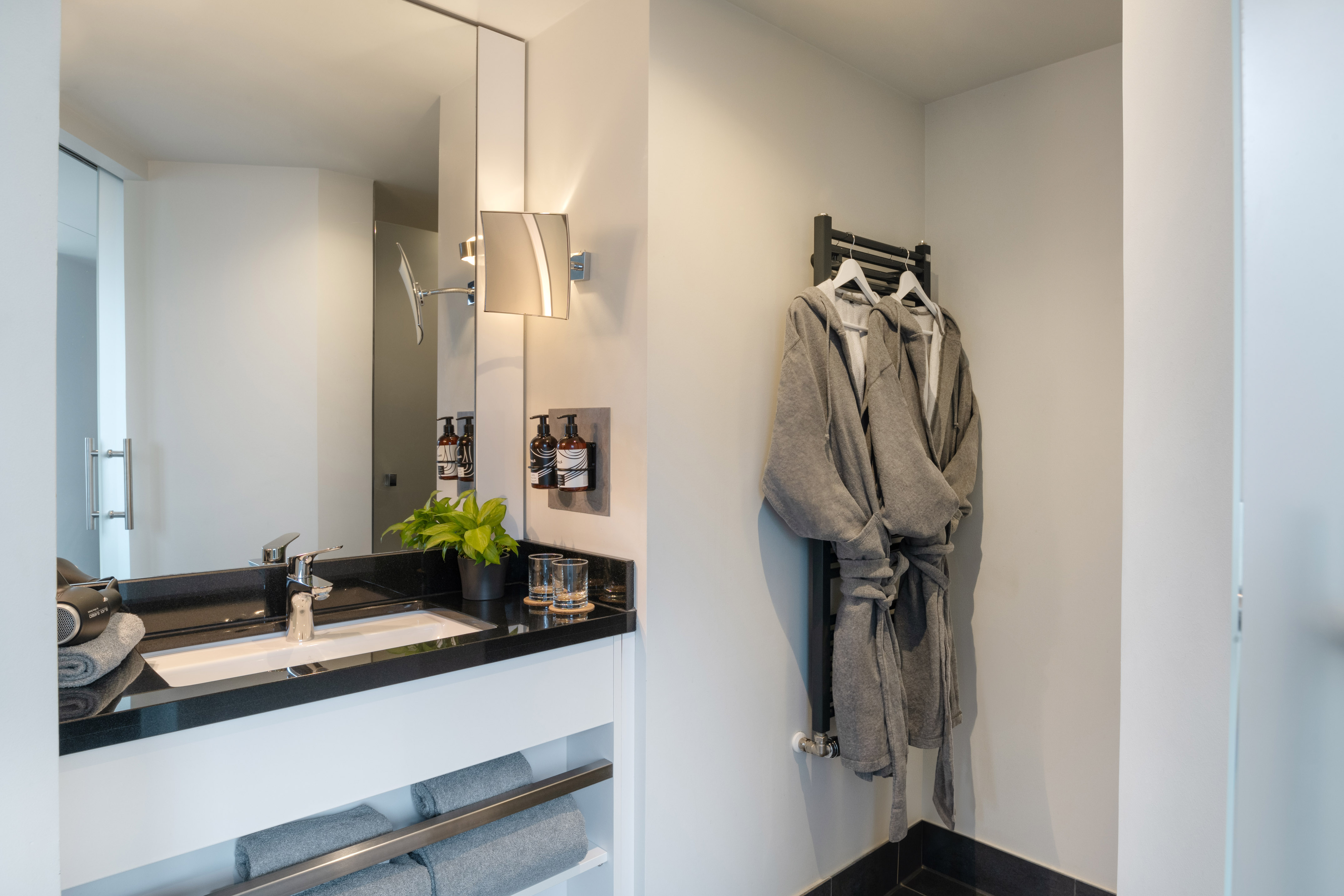 Modern hotel bathroom with black vanity, sink, mirror, bathrobes, and towels.