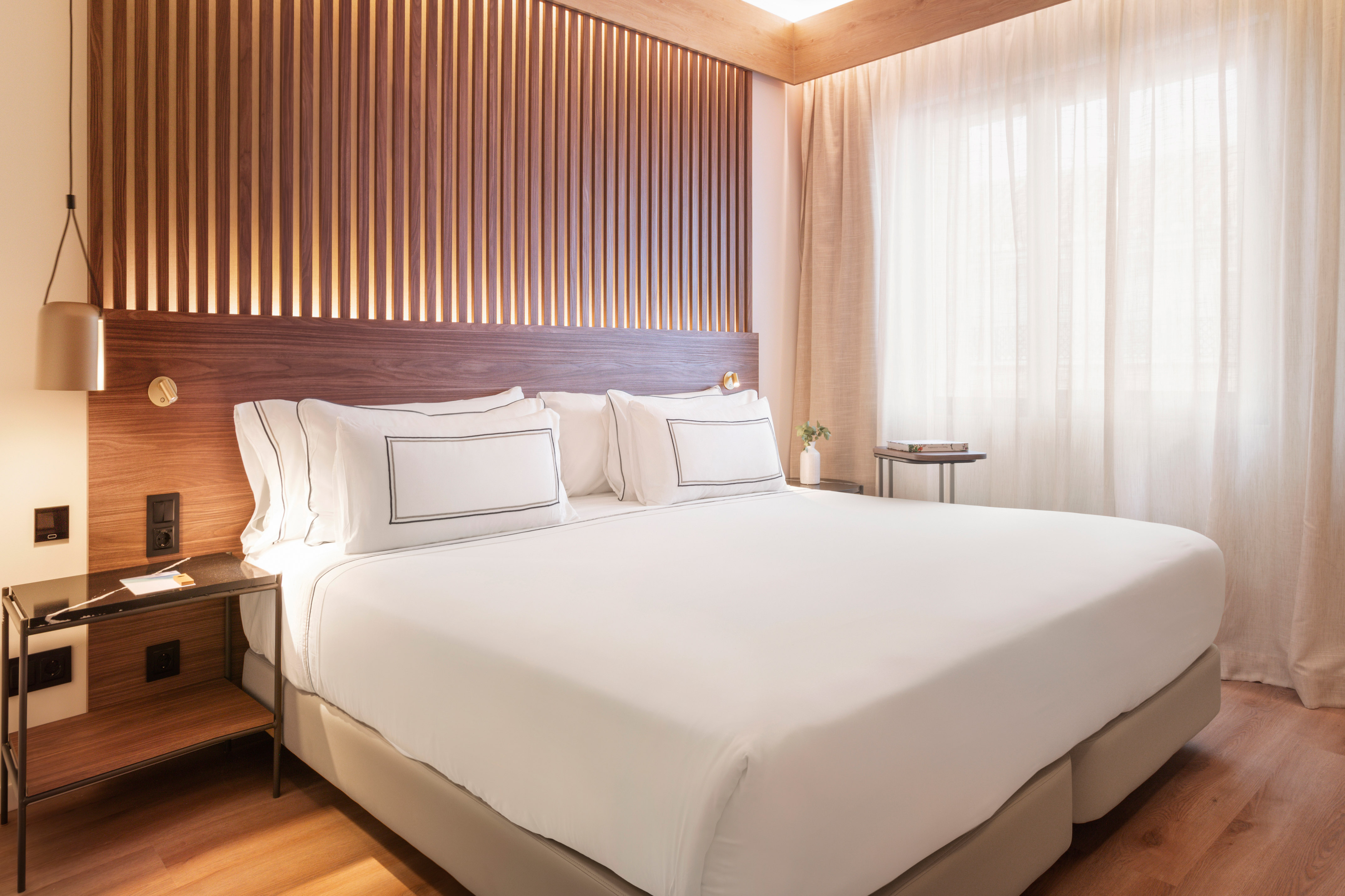 Modern bedroom with wooden slat headboard, white linens, and sheer curtains.