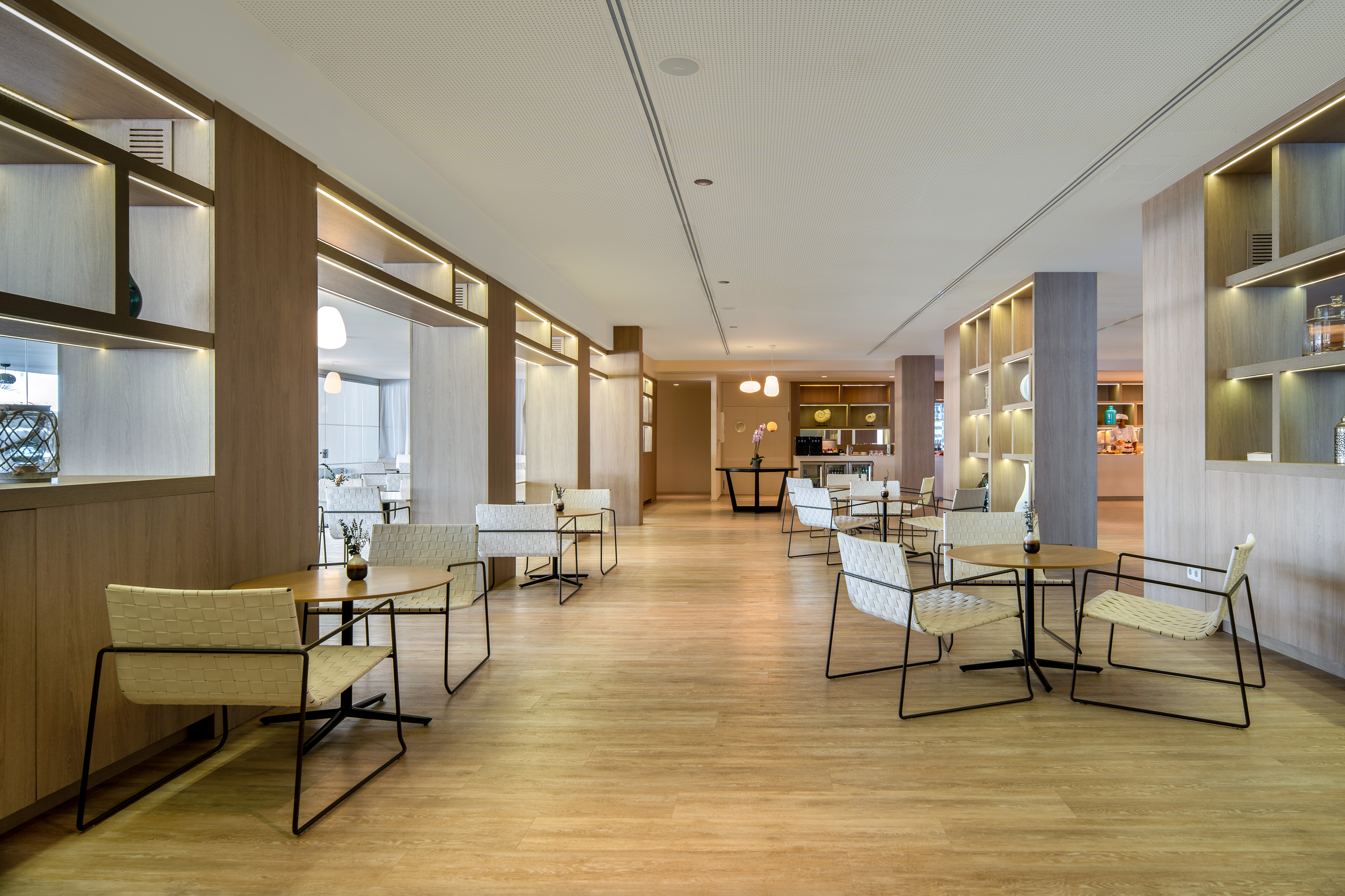 Modern cafe lounge with light wood floors, white chairs, and illuminated shelving.