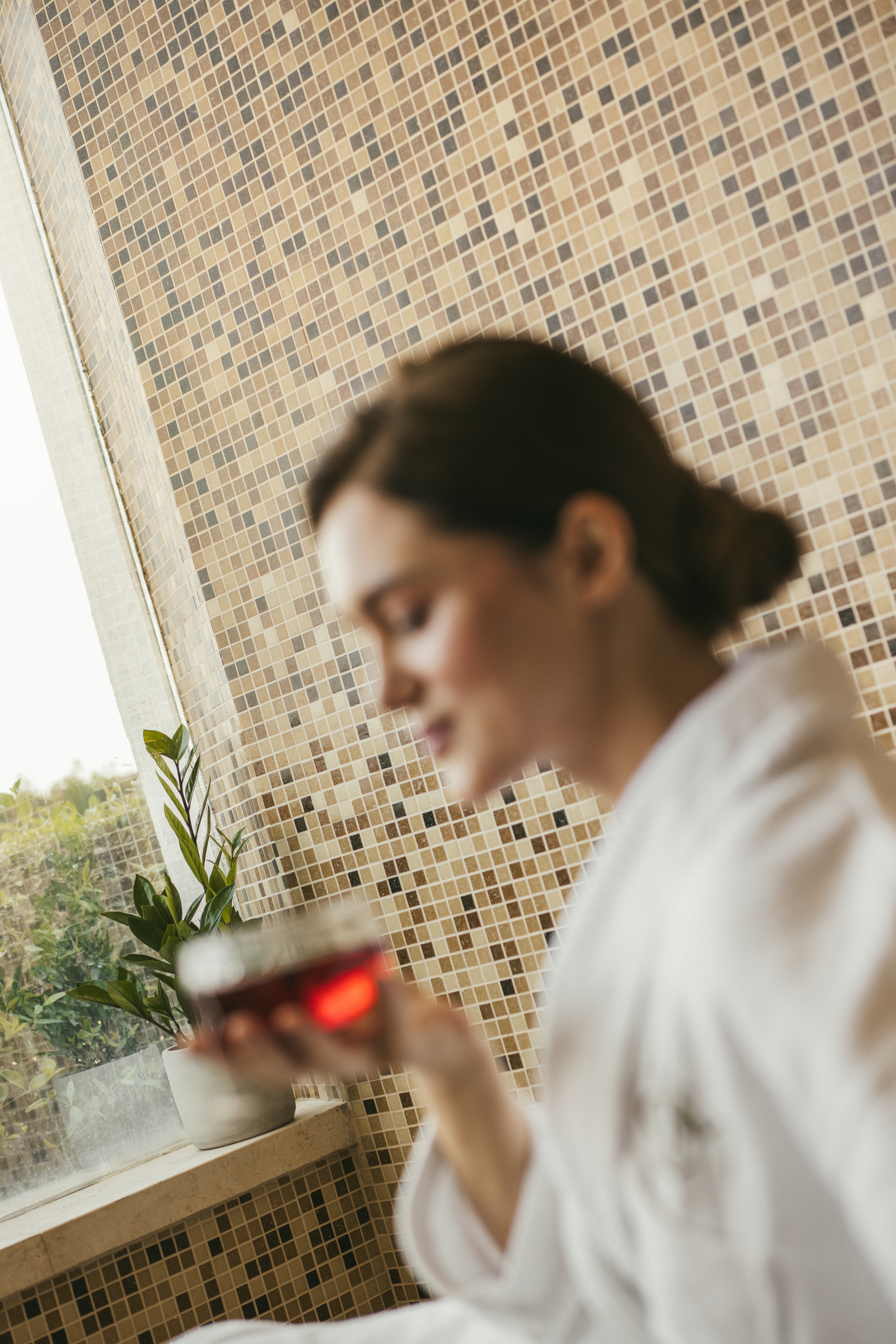 Woman in white bathrobe holding drink, mosaic wall, for spa relaxation.