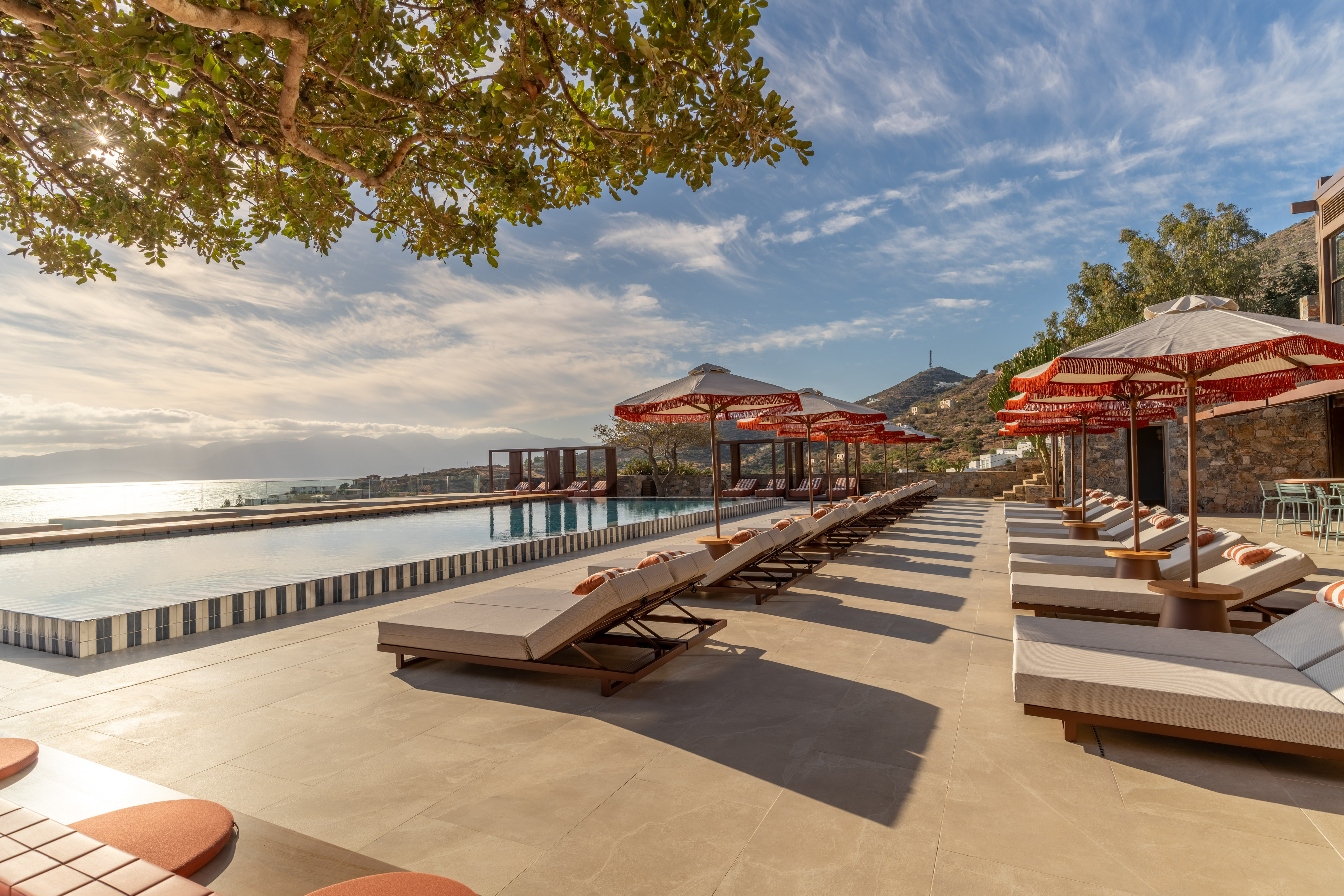 Luxury resort infinity pool with sea view, lounge chairs, and fringed sun umbrellas.