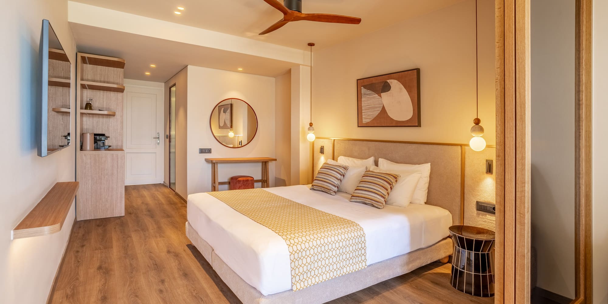 Modern hotel bedroom with king bed, wooden floor, beige walls, and ceiling fan.
